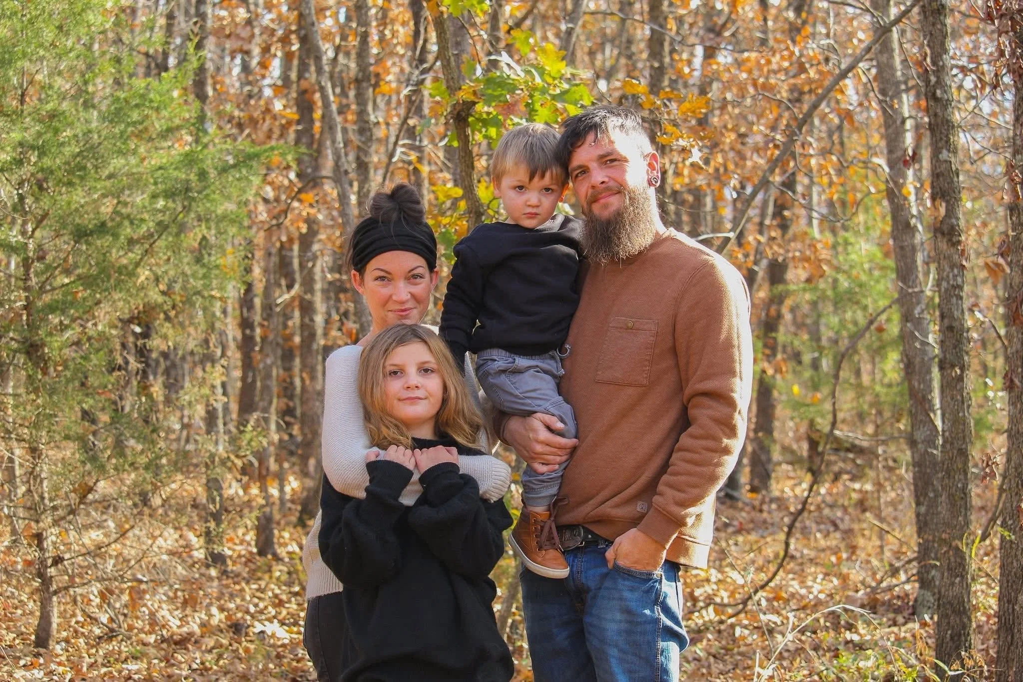 A family of five posing outdoors in a forest during autumn, with trees and fallen leaves in the background.