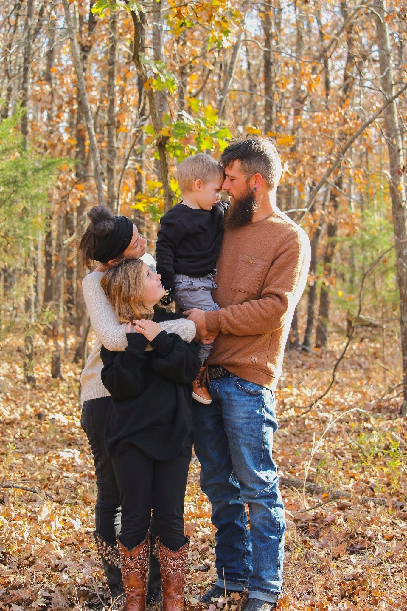 A man with a beard holds a young boy close to his forehead in a forest with autumn leaves, while a woman with a black headband and a young girl stand nearby in fall clothing, all surrounded by fallen leaves and trees.