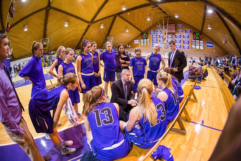 Girls' basketball team during a timeout, listening to coaches on the bench in an indoor gym, with scoreboard and banners in the background.