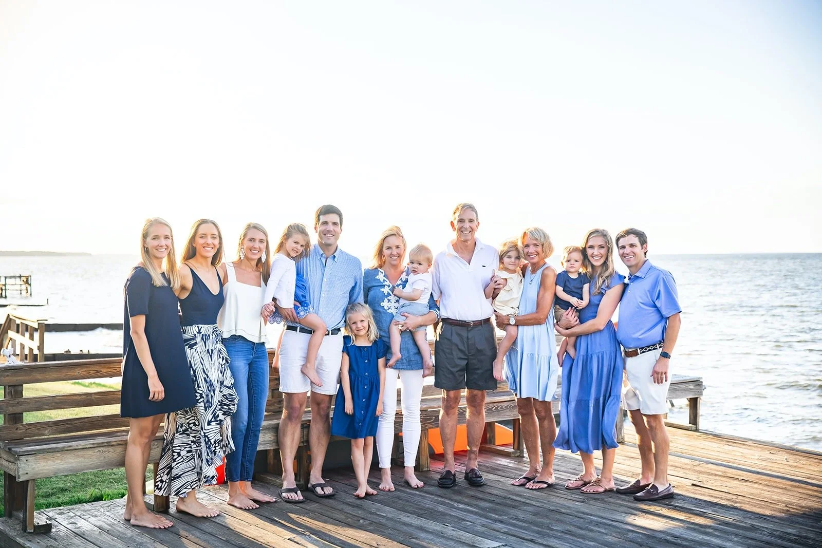 Family group standing on a wooden dock by the water, dressed in casual summer clothes, smiling at the camera during sunset.