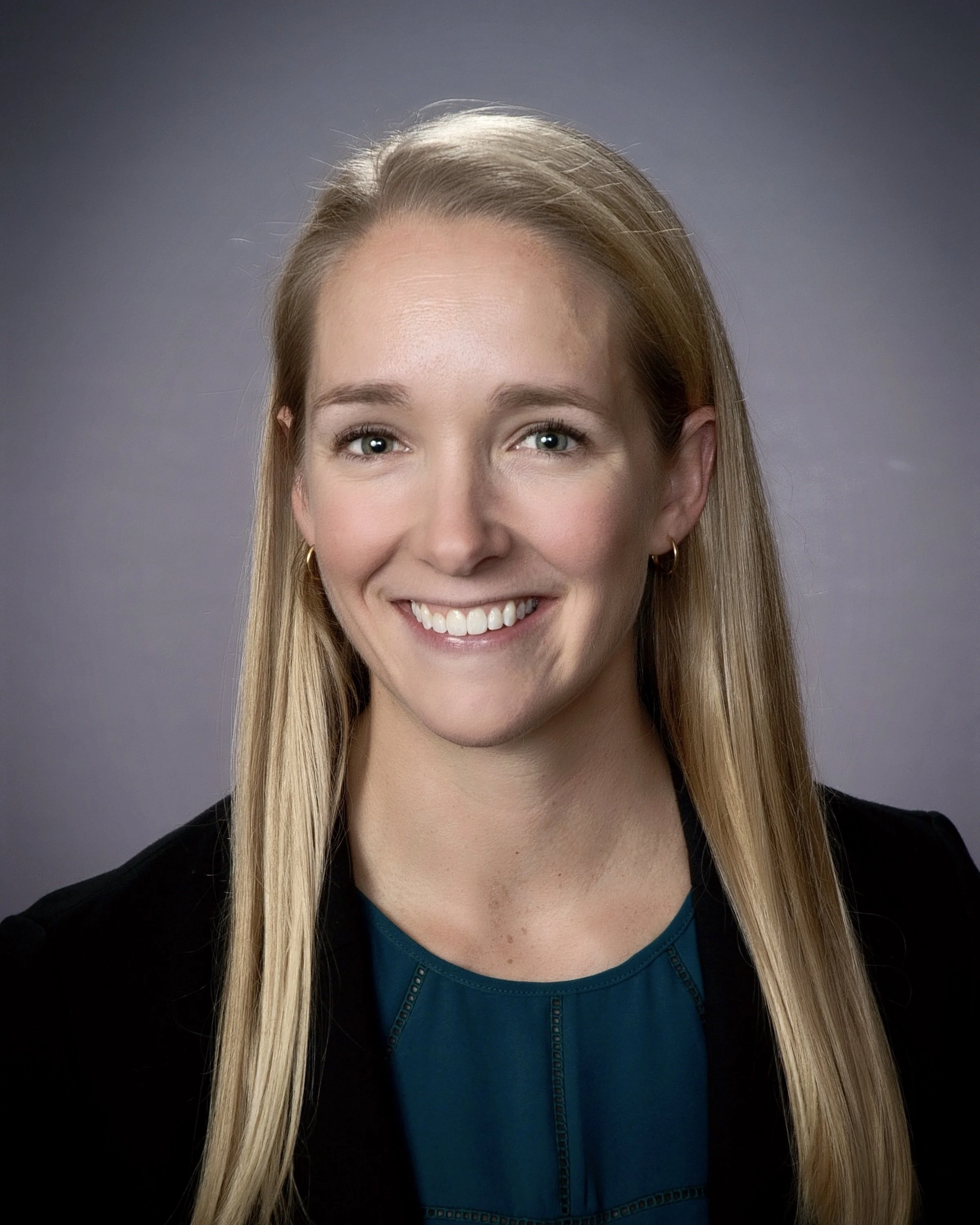 A smiling woman with long blonde hair, blue eyes, wearing a black blazer over a teal top, with a neutral gray background.