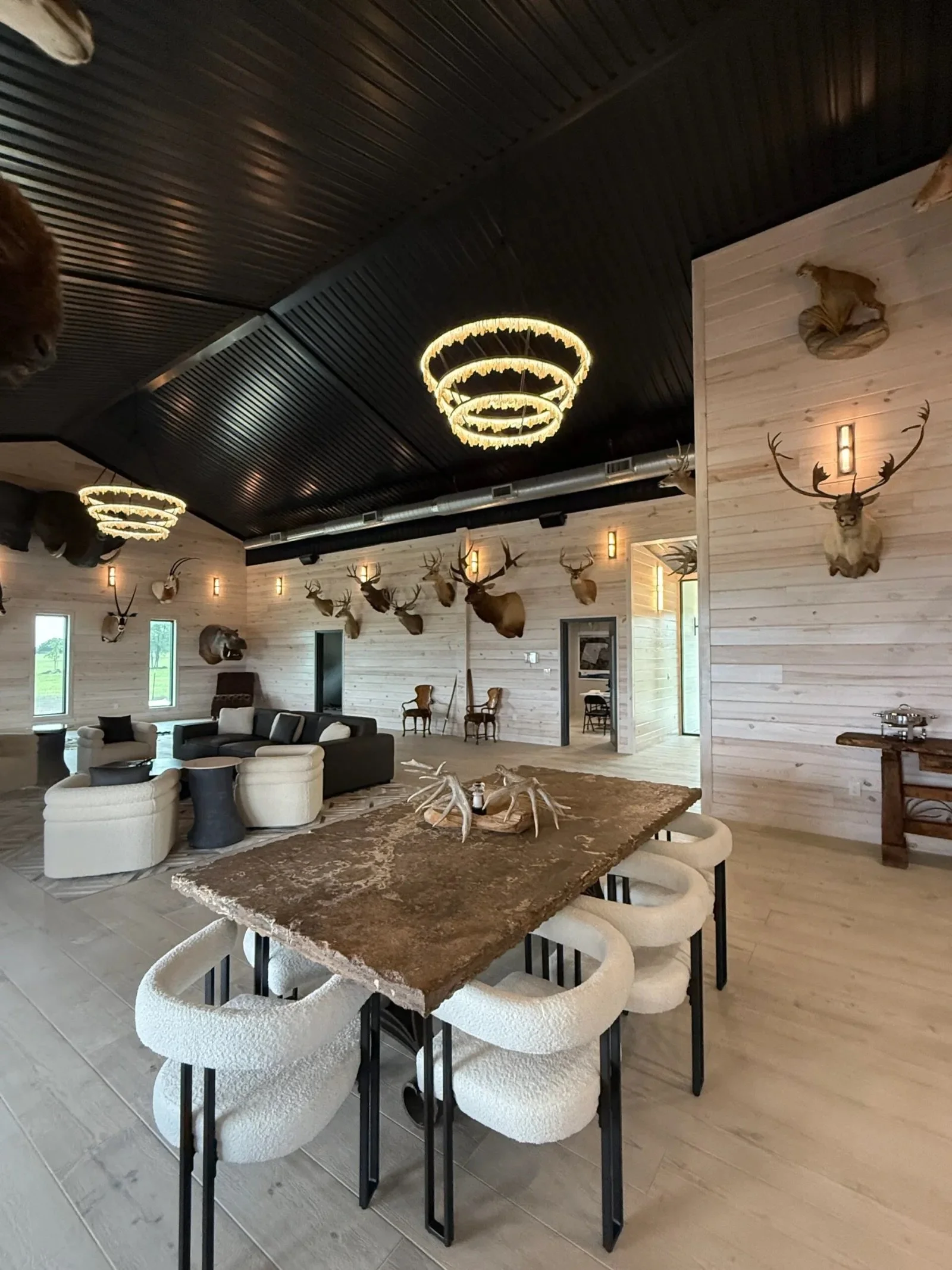 Interior of a room with wooden walls and mounted deer heads, modern chandeliers, and Victorian-style chairs.