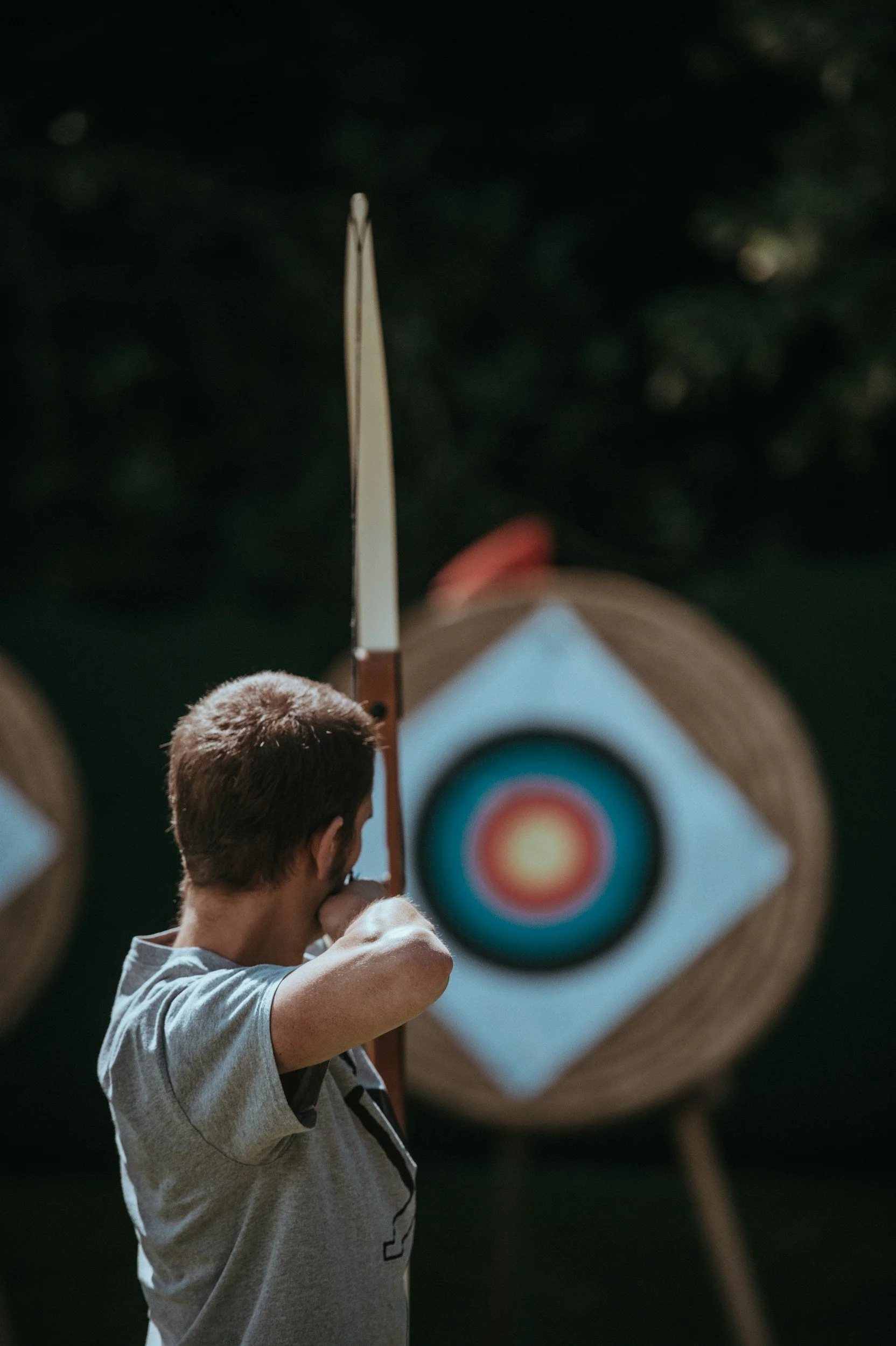 A young boy with brown hair and a gray shirt is preparing to throw an arrow at a target with colorful concentric circles, outdoors with a dark, blurred background and archery targets.