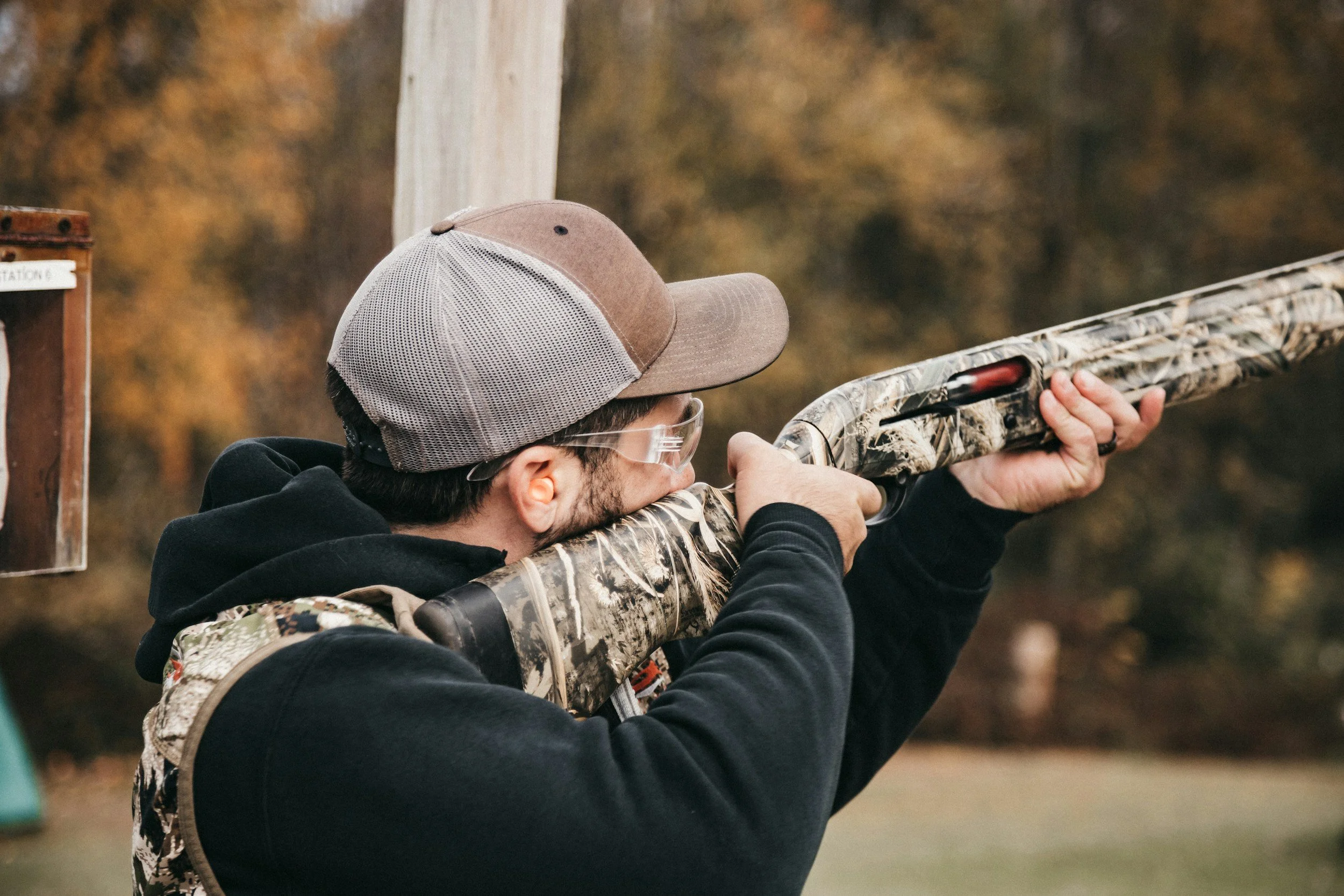 A man aiming a camouflage hunting rifle outdoors with autumn trees in the background.