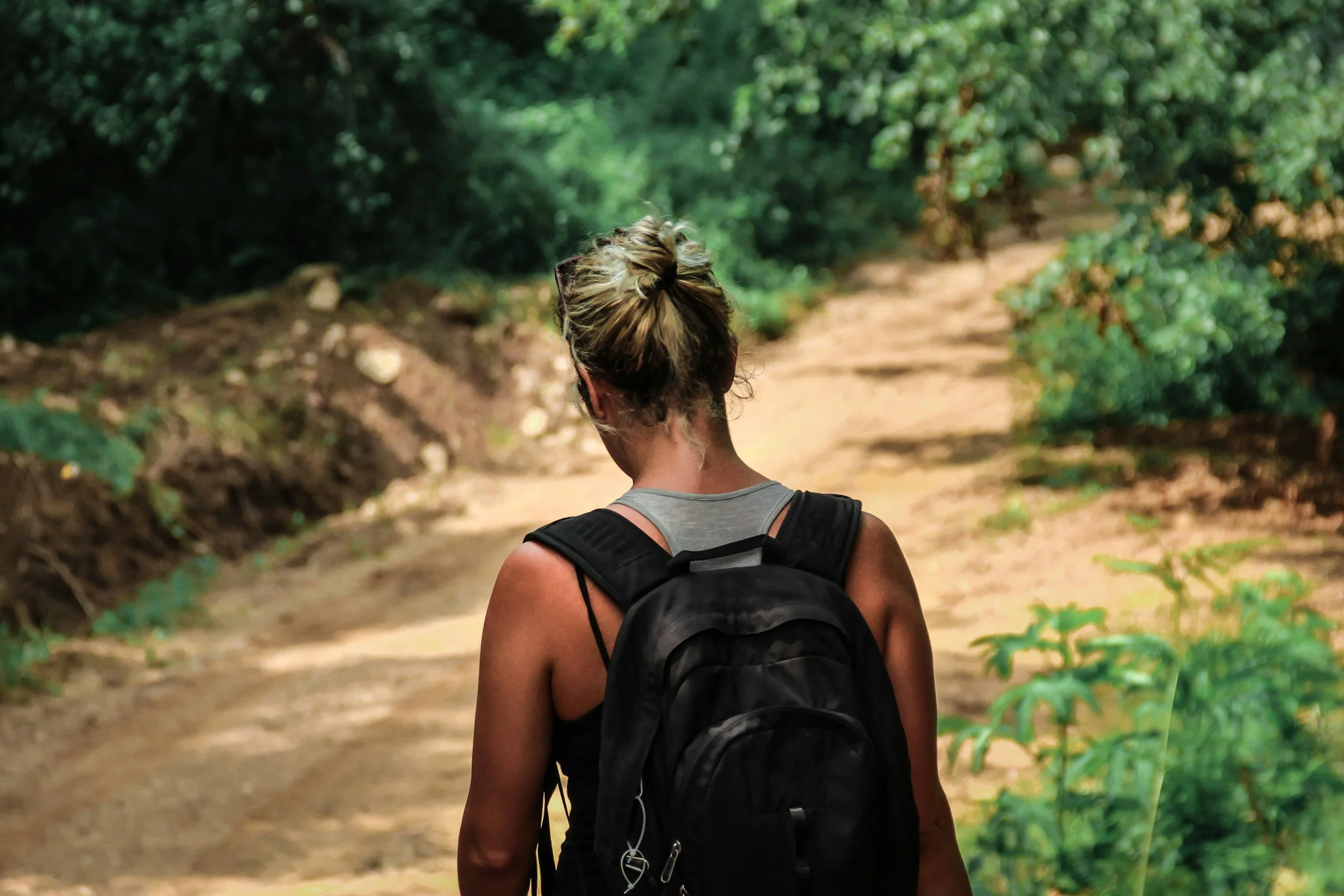 A woman with braided blonde hair wearing a gray tank top and black backpack walking on a dirt trail through a green forest during daytime. She is seen from behind.