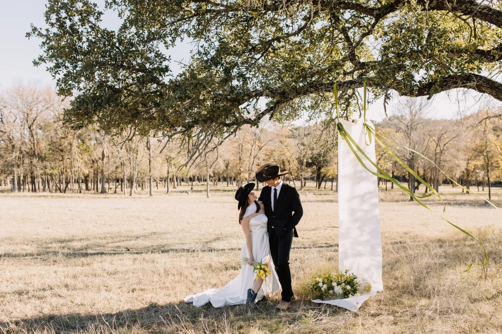A couple dressed in wedding attire standing under a tree in an open field, with a white wedding arch decorated with green ribbons and flowers beside them.