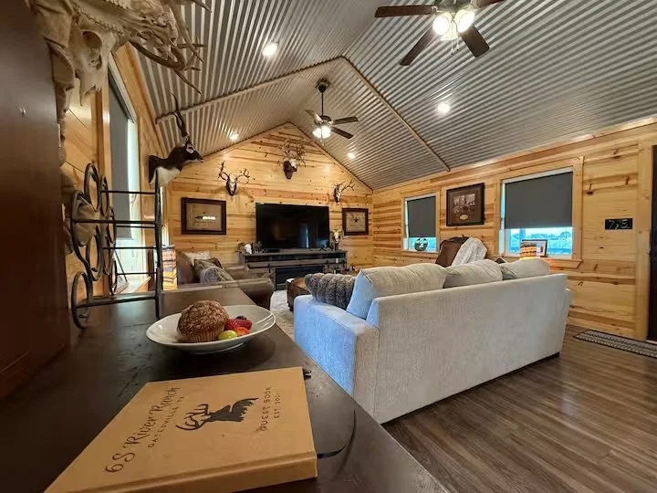 Rustic living room with wood-paneled walls and ceiling, decorated with mounted antlers, a large flat-screen TV, framed artwork, and windows with blinds, featuring a white upholstered sofa and a dark wooden coffee table.