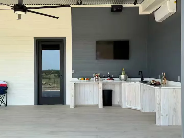Outdoor kitchen area with a dark gray wall, white cabinets, a mounted flat-screen TV, a small black trash bin, and a countertop with bottles, utensils, and food items. There is a door with glass and a black door handle to the left, a ceiling fan, and an air conditioning unit on the wall.