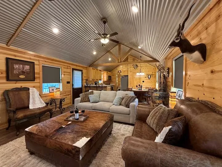 Living room and dining area in a rustic cabin with wooden walls and ceiling, furniture including a white sofa, leather armchair, and wooden coffee table, with a mounted deer head and a taxidermy goat on the walls, and a ceiling fan.