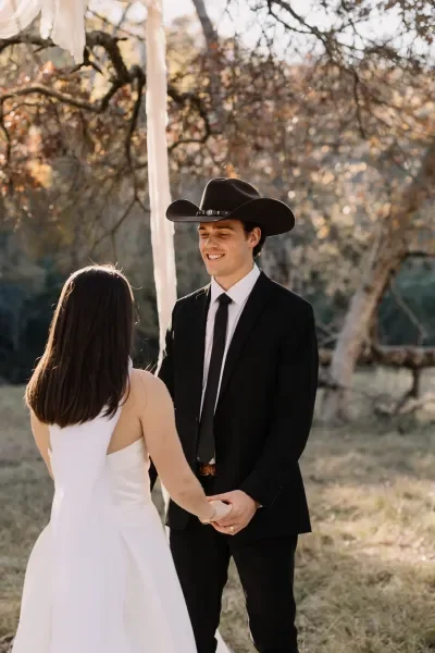 A young man in a black suit and cowboy hat holding hands with a woman in a white dress outdoors during daytime, with trees in the background.
