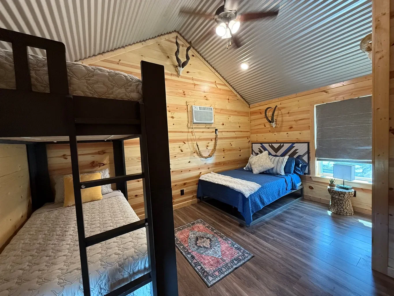 Rustic bedroom with wooden walls, a bunk bed with mattresses, a full bed with blue bedding and pillows, a small rug, a window with a blind, a nightstand with a lamp, and mounted animal skulls and horns on the wall.