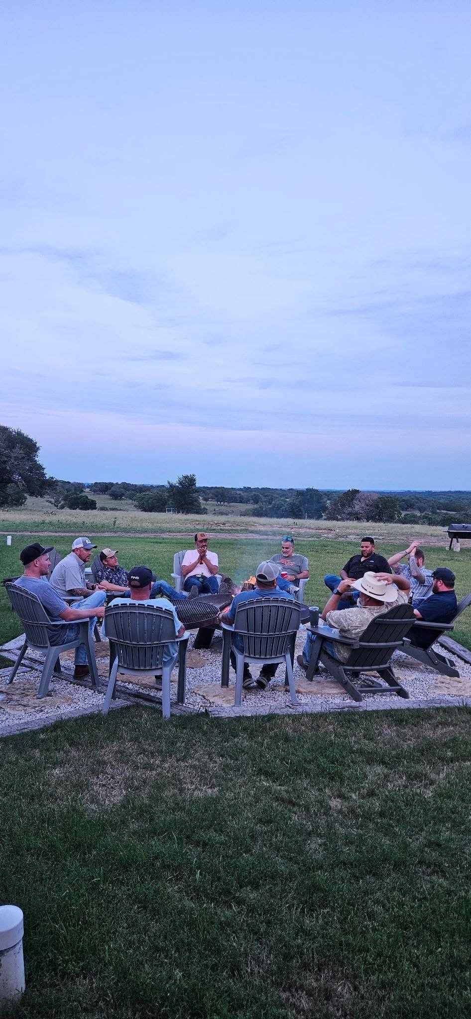 A group of people sitting around a fire pit outdoors during dusk on a grassy lawn, with open fields and trees in the background.