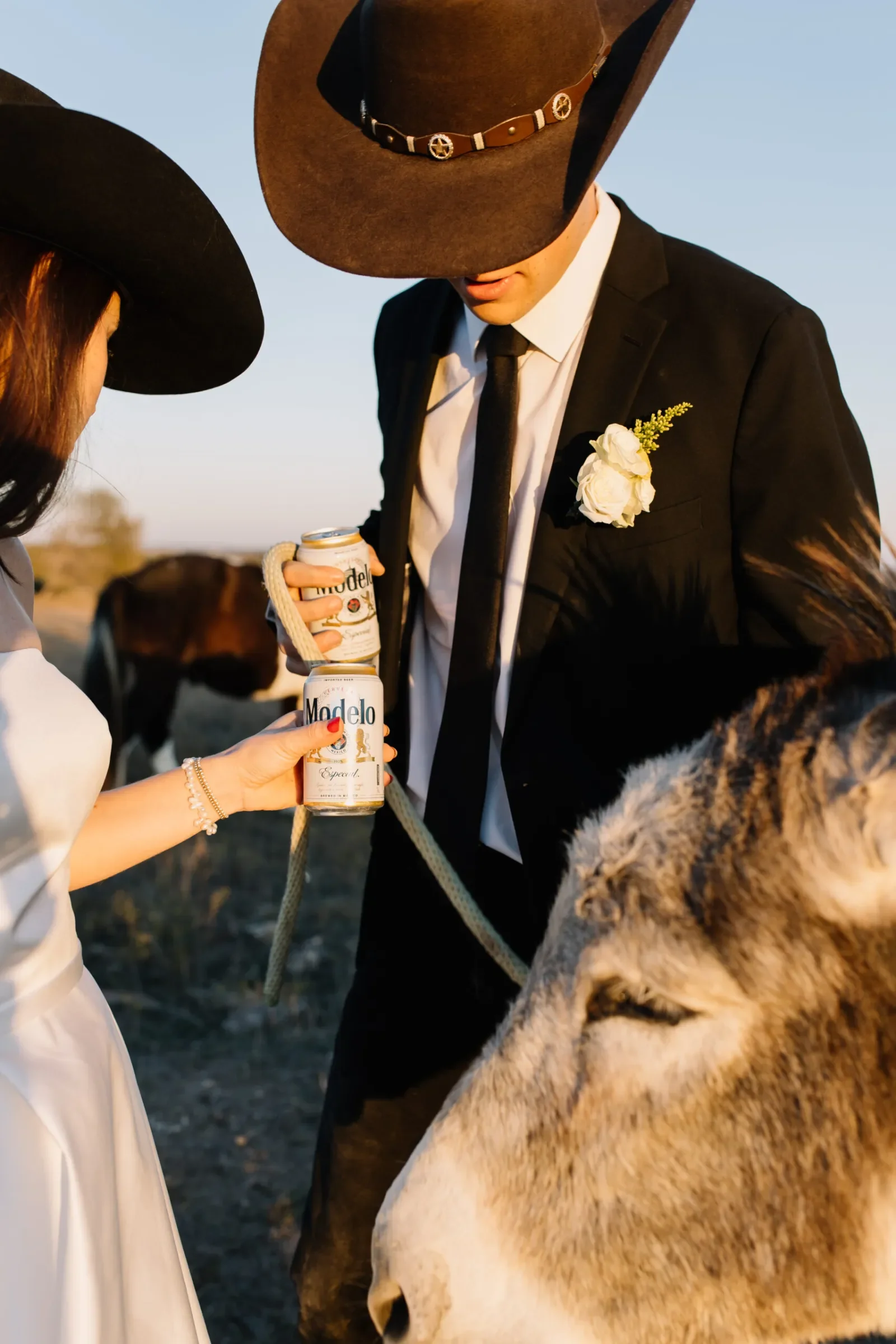 A man dressed in a black suit with a white boutonniere and a woman in a white dress both wearing black hats with wide brims are sharing a moment outdoors as they hold up cans of Modelo beer during sunset.