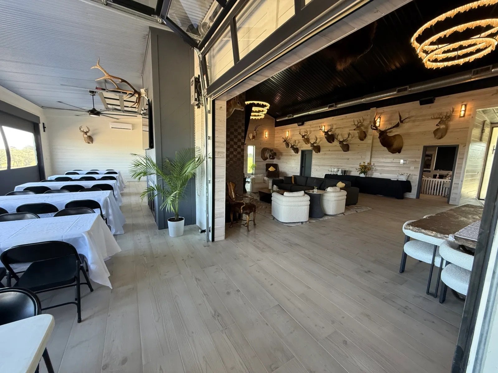 Interior of a rustic-style event space or restaurant with white-washed wooden walls and floors, decorated with mounted deer heads, chandeliers, and a variety of seating areas including tables with white tablecloths, lounge chairs, and a bar area.