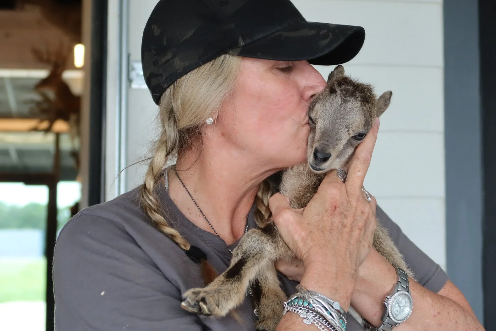 A woman with blonde hair in braids, wearing a black camouflage cap and a gray shirt, is kissing a baby goat on the head while holding it close to her face.