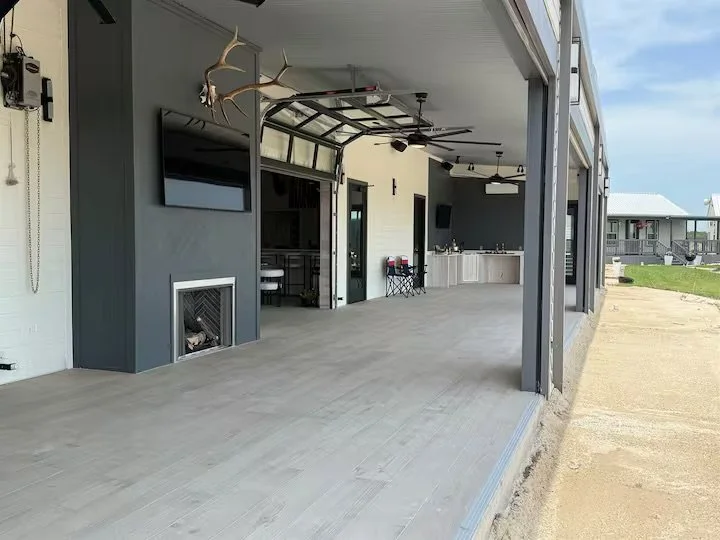 Large open outdoor patio area with gray flooring, features a wall-mounted TV, a fireplace, a hanging deer head, and a partial view of an outdoor kitchenette with counters and cabinets. The space is attached to a house with neighboring houses visible in the background.