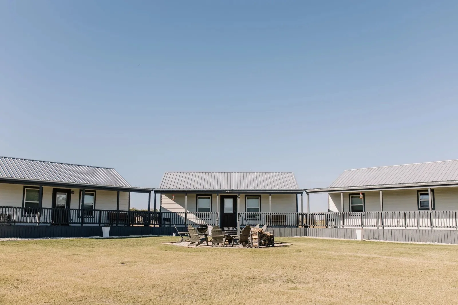 Three white houses with black trim and metal roofs, set against a clear blue sky. In the foreground, there is a grassy lawn with a circle of patio chairs and a barbecue grill.