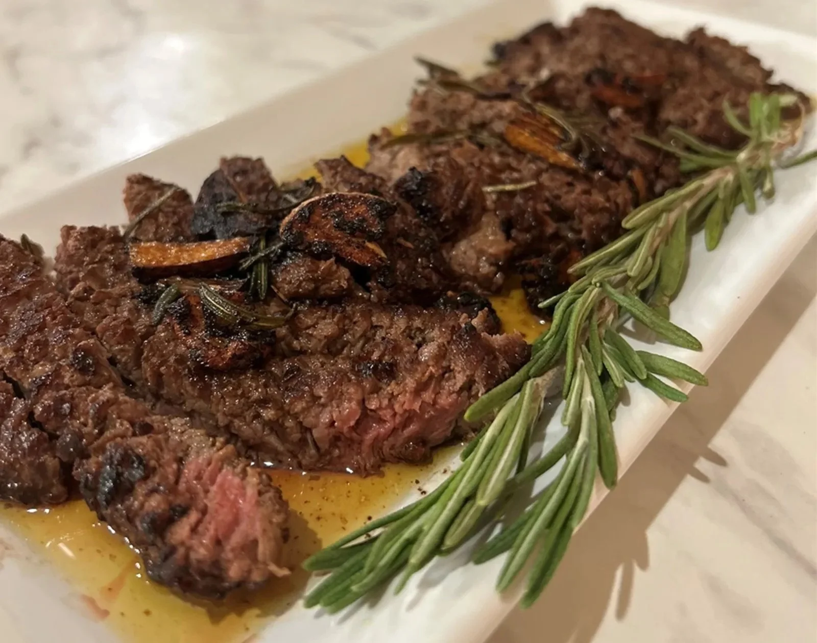 Close-up of cooked beef meatloaf topped with roasted garlic and rosemary, garnished with a sprig of fresh rosemary on a white plate.