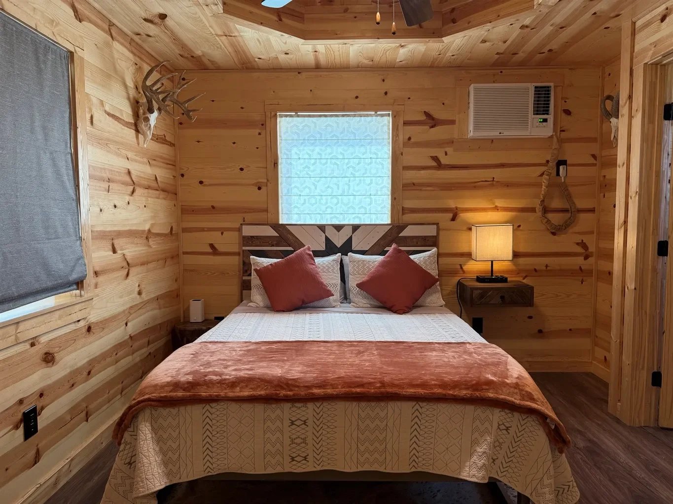 A cozy bedroom with wooden panel walls and ceiling, featuring a double bed with white, beige, and rust-colored bedding, two rust-colored pillows, and a headboard with a geometric wooden pattern. There is a window with a patterned curtain behind the bed, a nightstand with a lamp on the right side, a small speaker on the left side, and mounted antlers on the left wall. An air conditioning unit is on the wall above the nightstand.