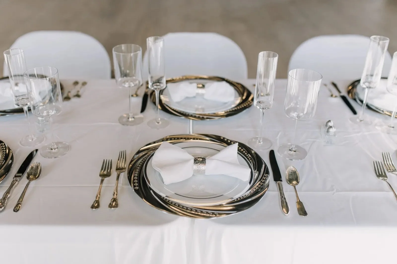 Elegant table setting with white tablecloth, black and silver plates, white napkin, and multiple wine glasses, chairs in the background.
