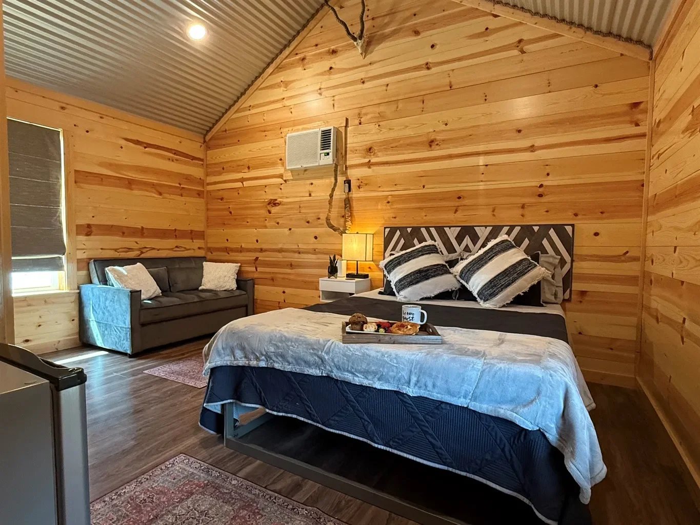 A cozy bedroom with wood-paneled walls and a sloped ceiling. It features a bed with black and white striped pillows, a bedside table with a lamp, a gray loveseat with pillows, and a tray with breakfast and a coffee mug on the bed.