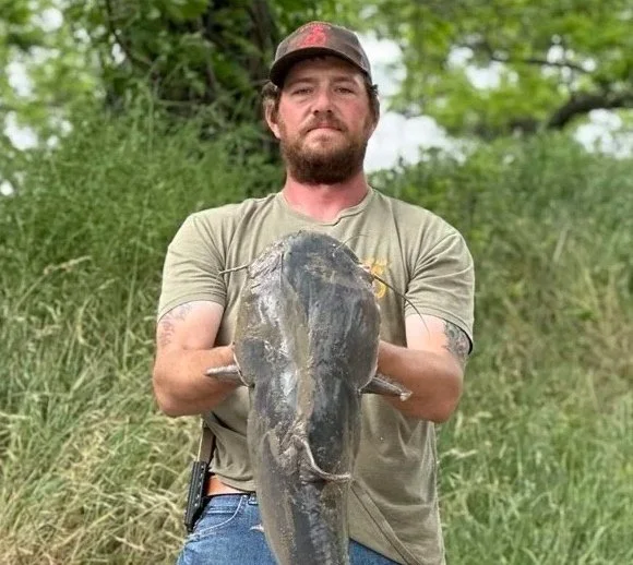 Man holding a large catfish outdoors in a grassy area with trees in the background.