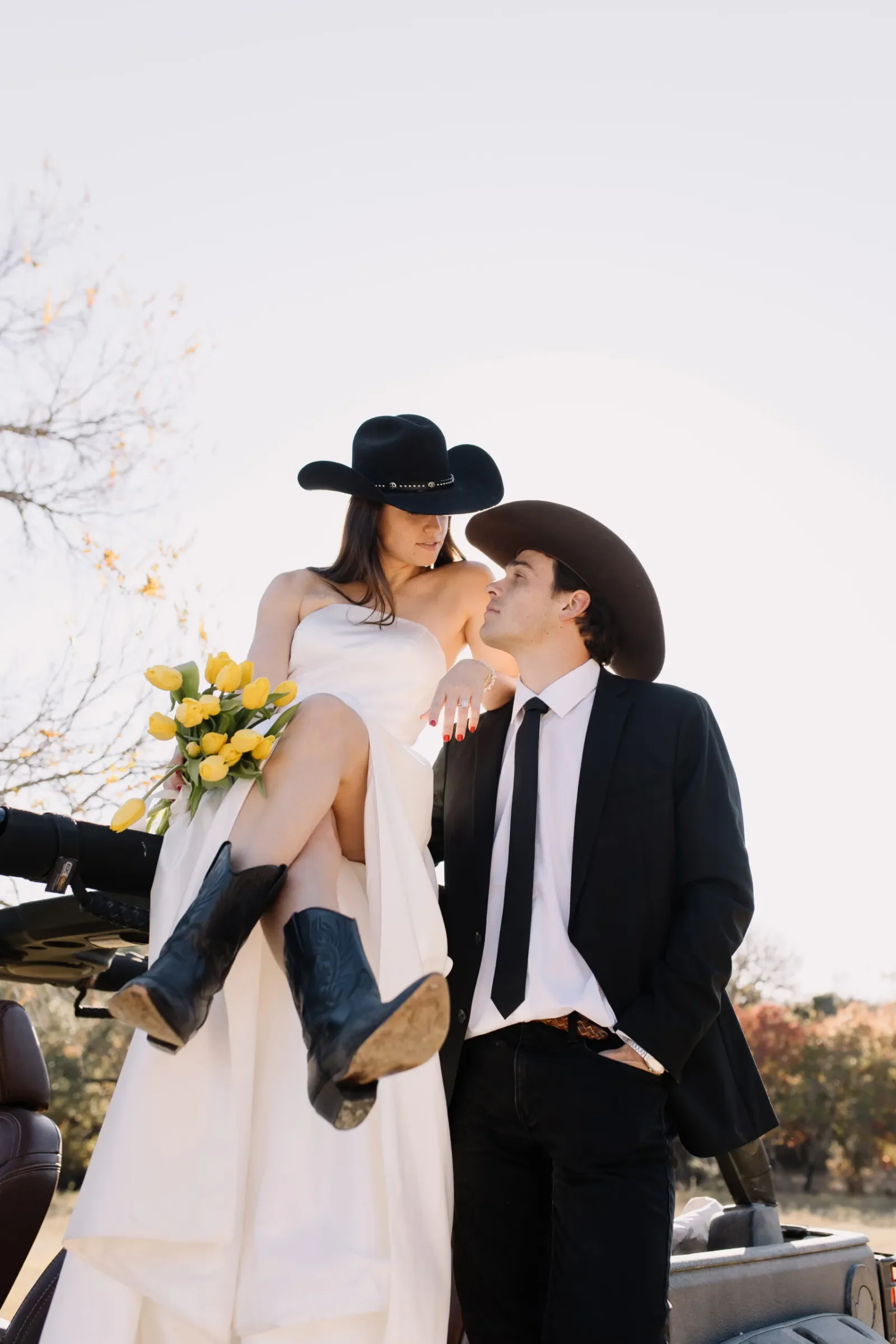 A woman in a white dress and black cowboy hat holding a bouquet of yellow tulips is sitting on a man’s lap. The man, dressed in a suit with a black hat, is standing and looking at her. They are outdoors with trees and clear sky in the background.