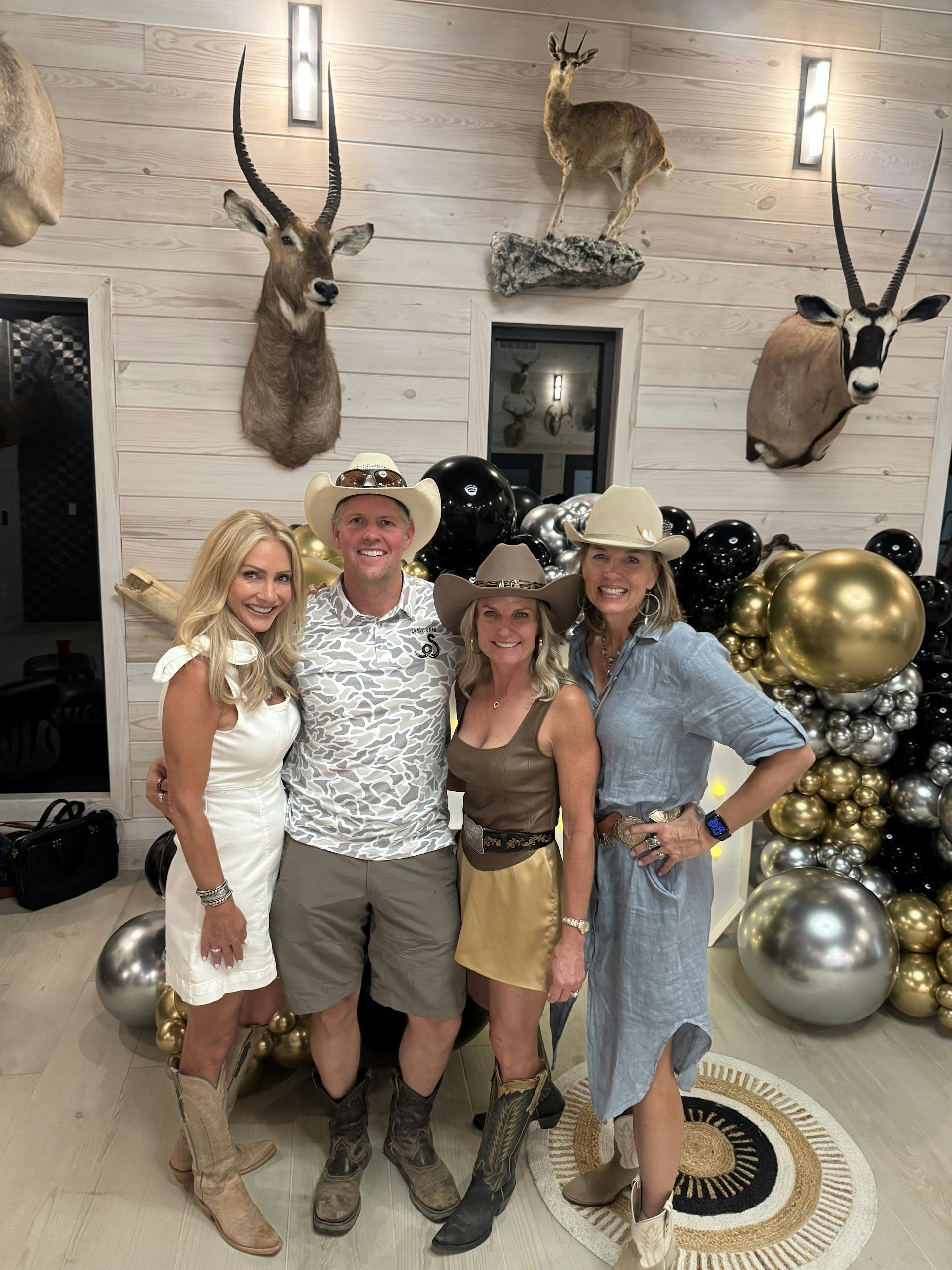 Four people dressed in cowboy or country-themed outfits, standing in front of a wall decorated with mounted animal heads, including an antelope, a small deer, and another antelope. There are also black, gold, and silver balloons arranged around them.