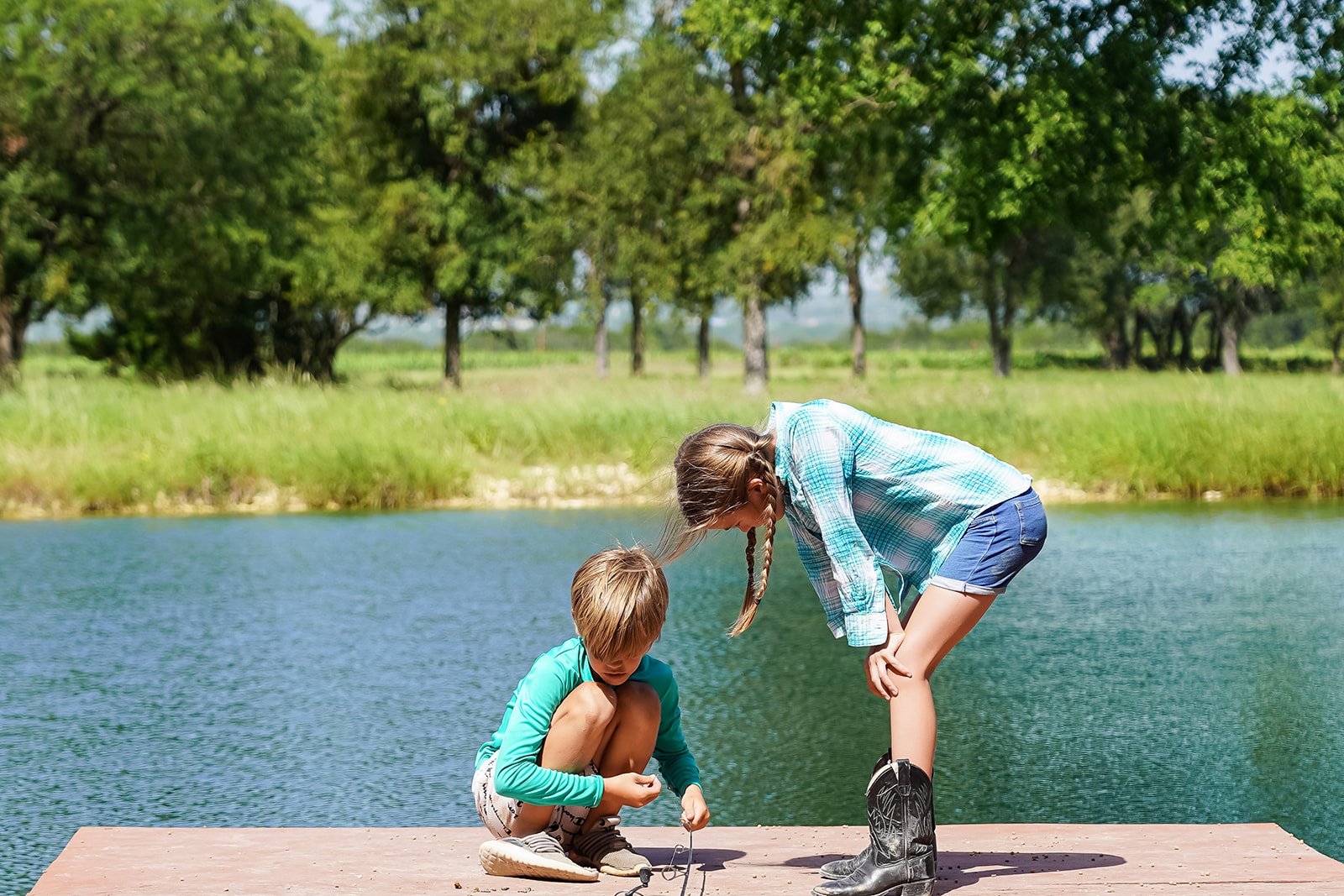 Two children, a girl and a boy, are crouched on a wooden dock by a lake, fishing and looking at their fishing lines, with trees and greenery in the background on a sunny day.