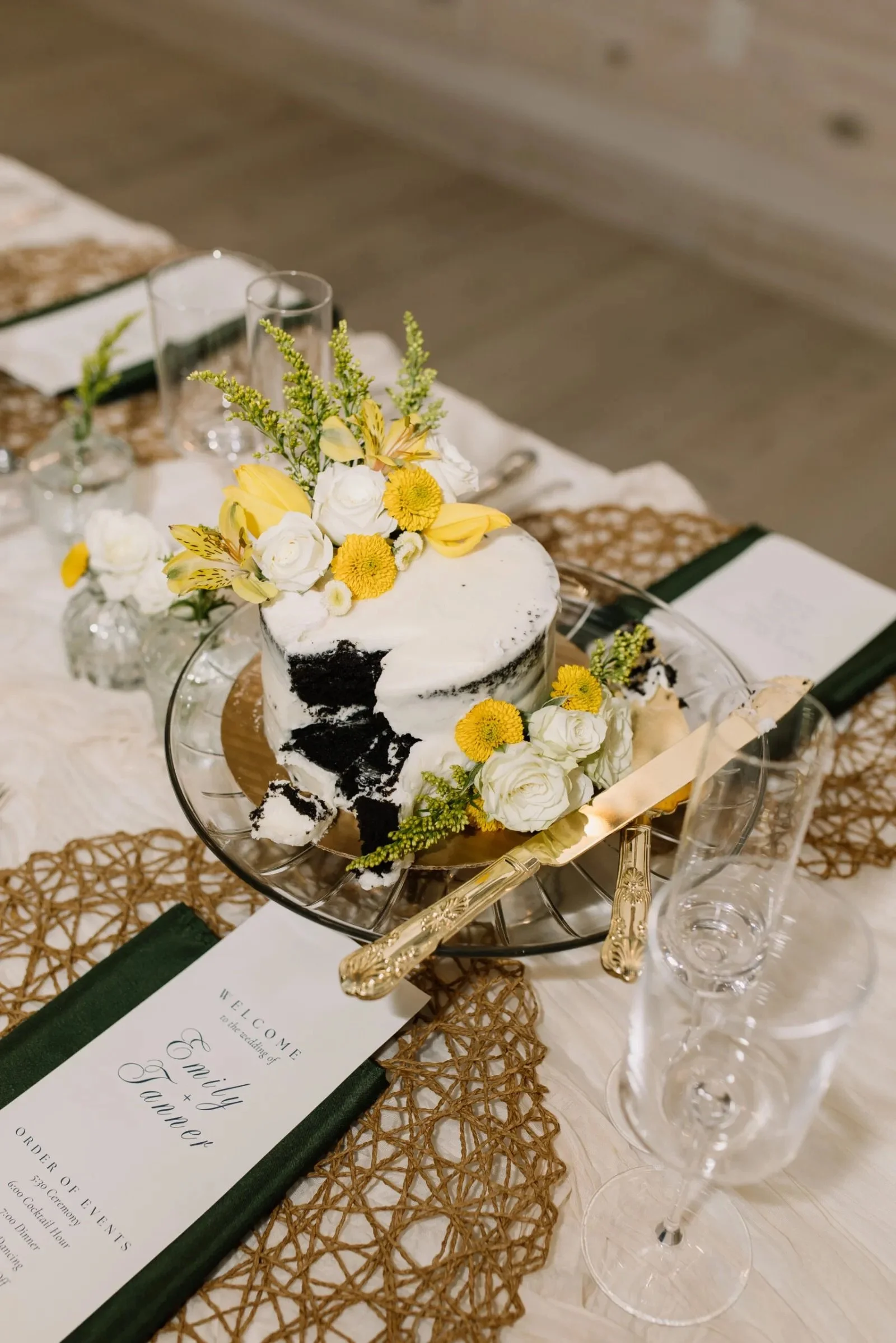 A decorated wedding reception table featuring a two-tiered white and black marbled cake with yellow and white flowers, surrounded by champagne glasses, a menu, and a decorative runner on a rustic table.