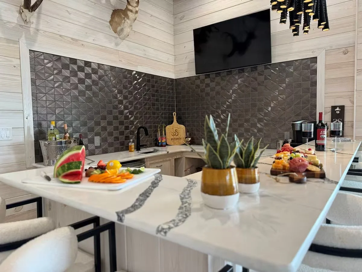 Modern kitchen with a white marble island, black textured backsplash, mounted TV, and various fruits and plants on the counter and island.