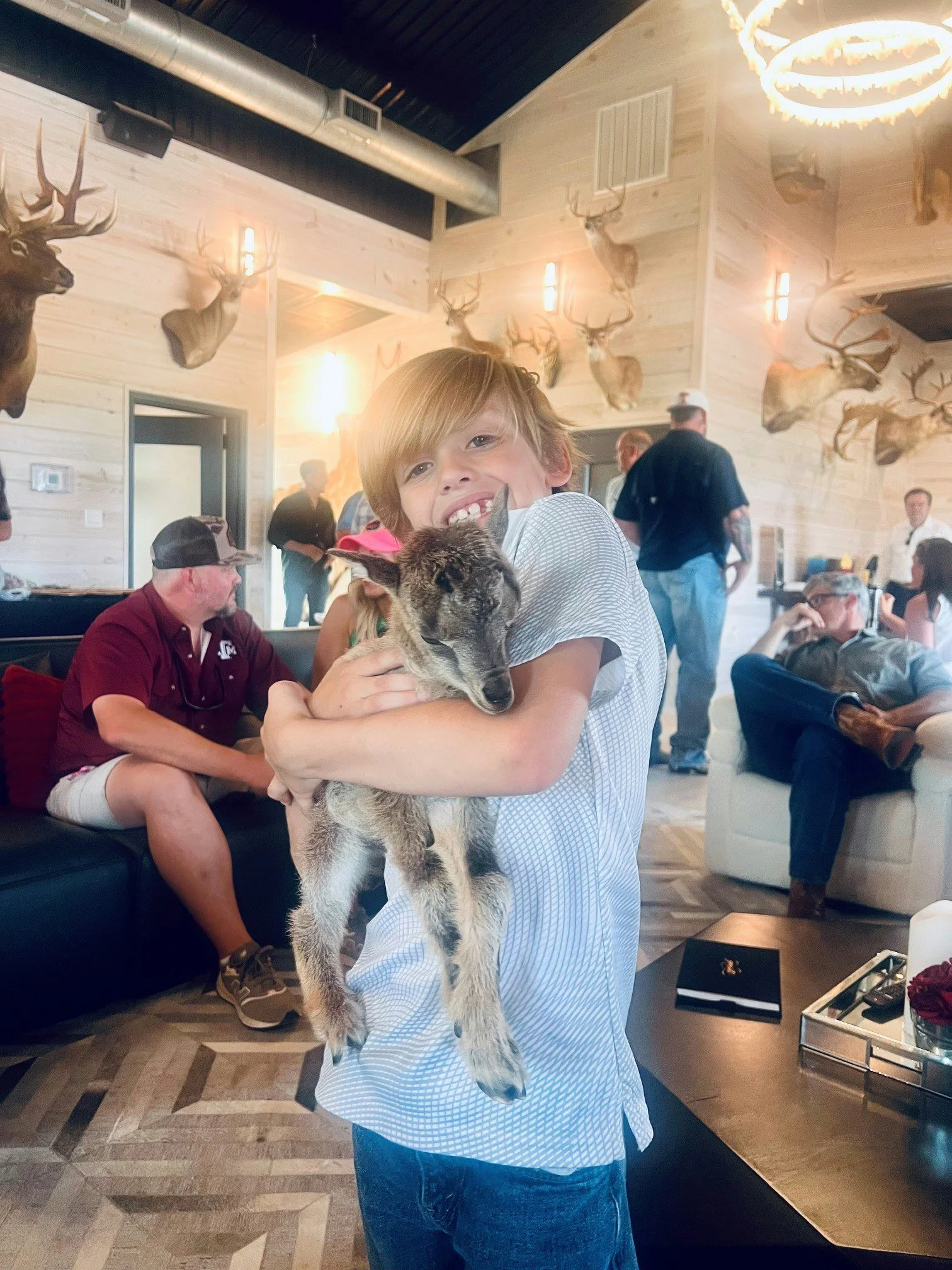 A young boy with brown hair and a gap-tooth smile is holding a small goat inside a rustic restaurant or lodge decorated with mounted elk and deer heads on the walls. Several adults are seated and standing in the background.