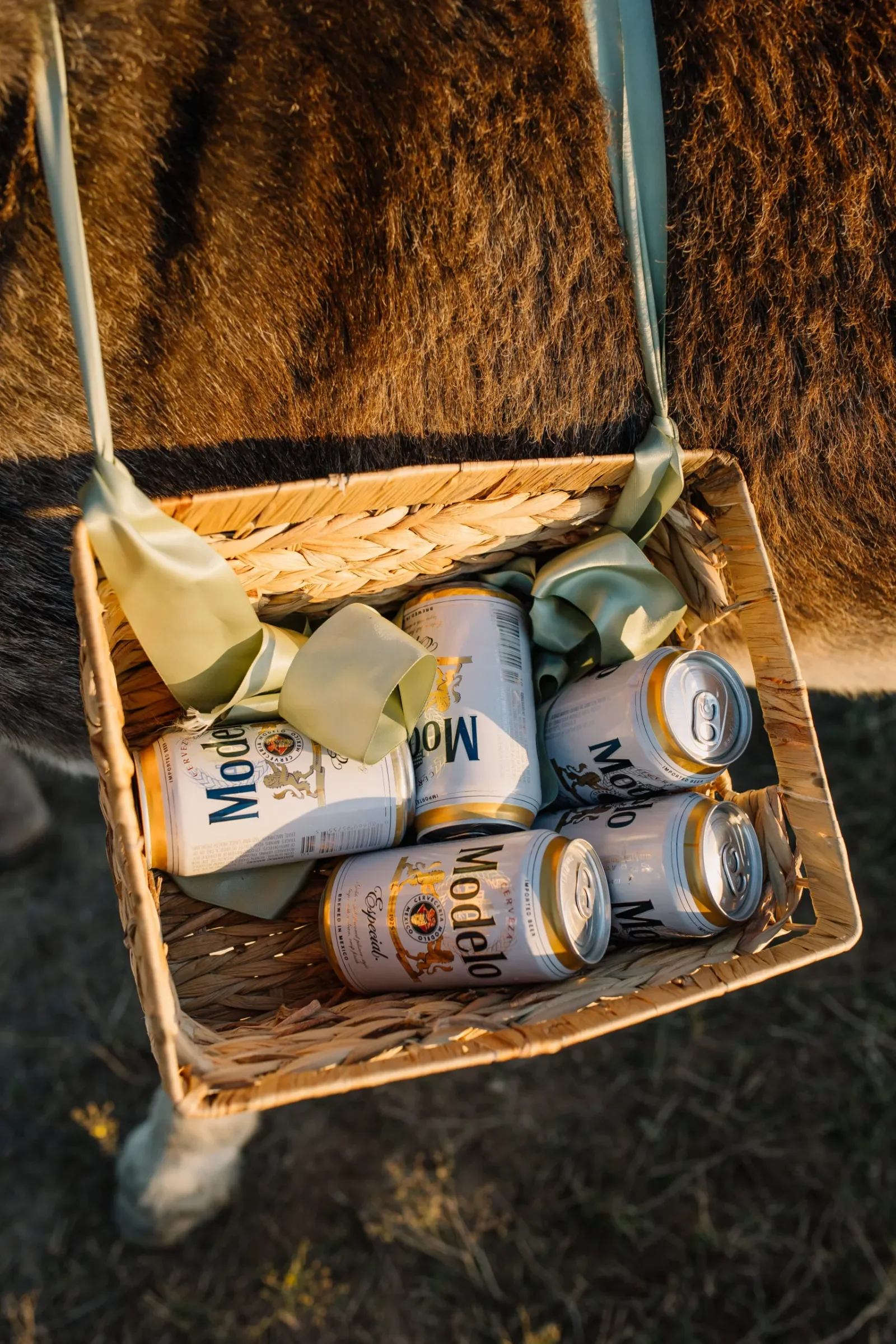 A woven basket filled with empty beer cans placed outside on dirt with sparse grass.