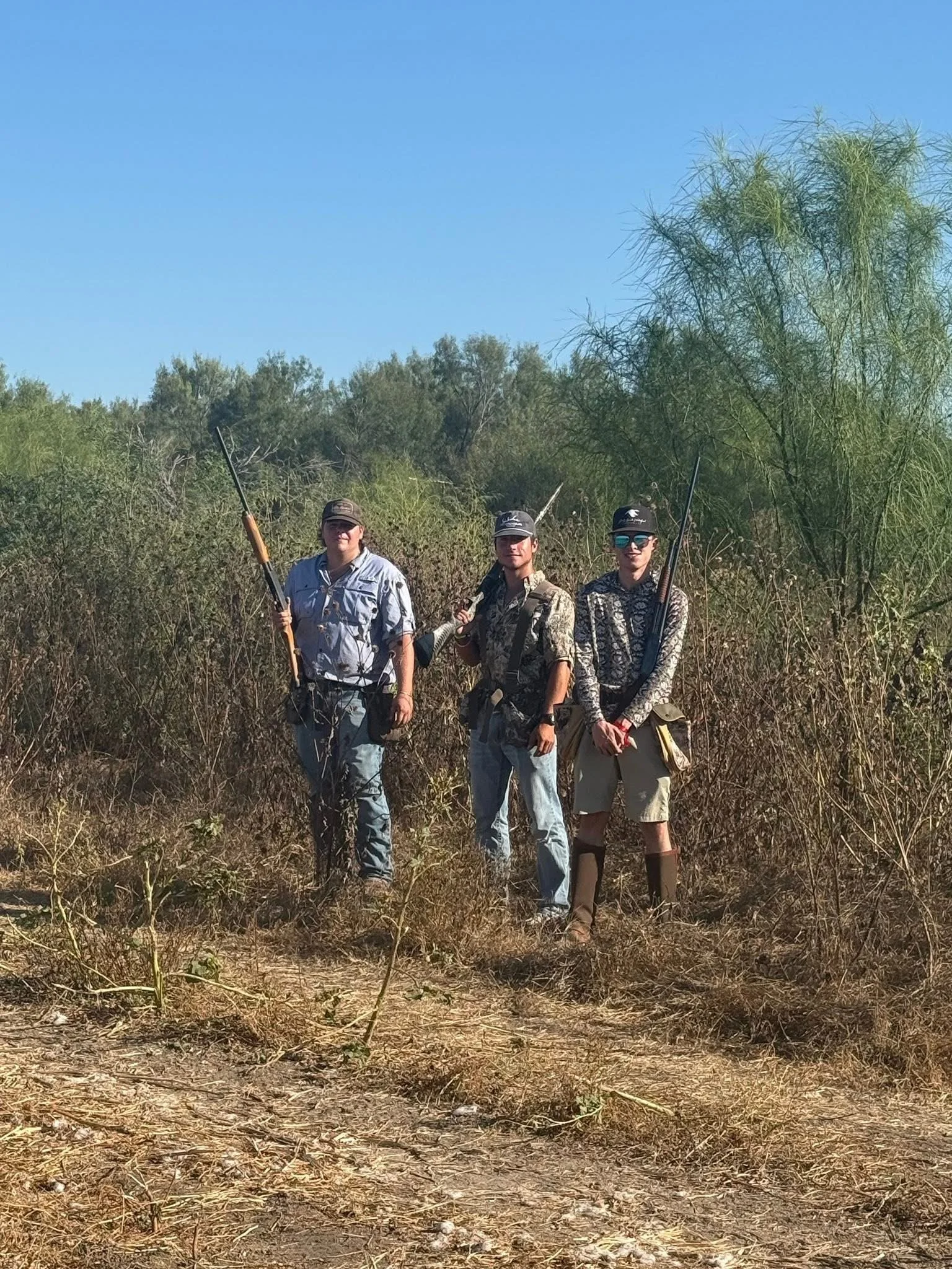 Three people standing in a dry, grassy, outdoor area with bushes and trees behind them, holding rifles.