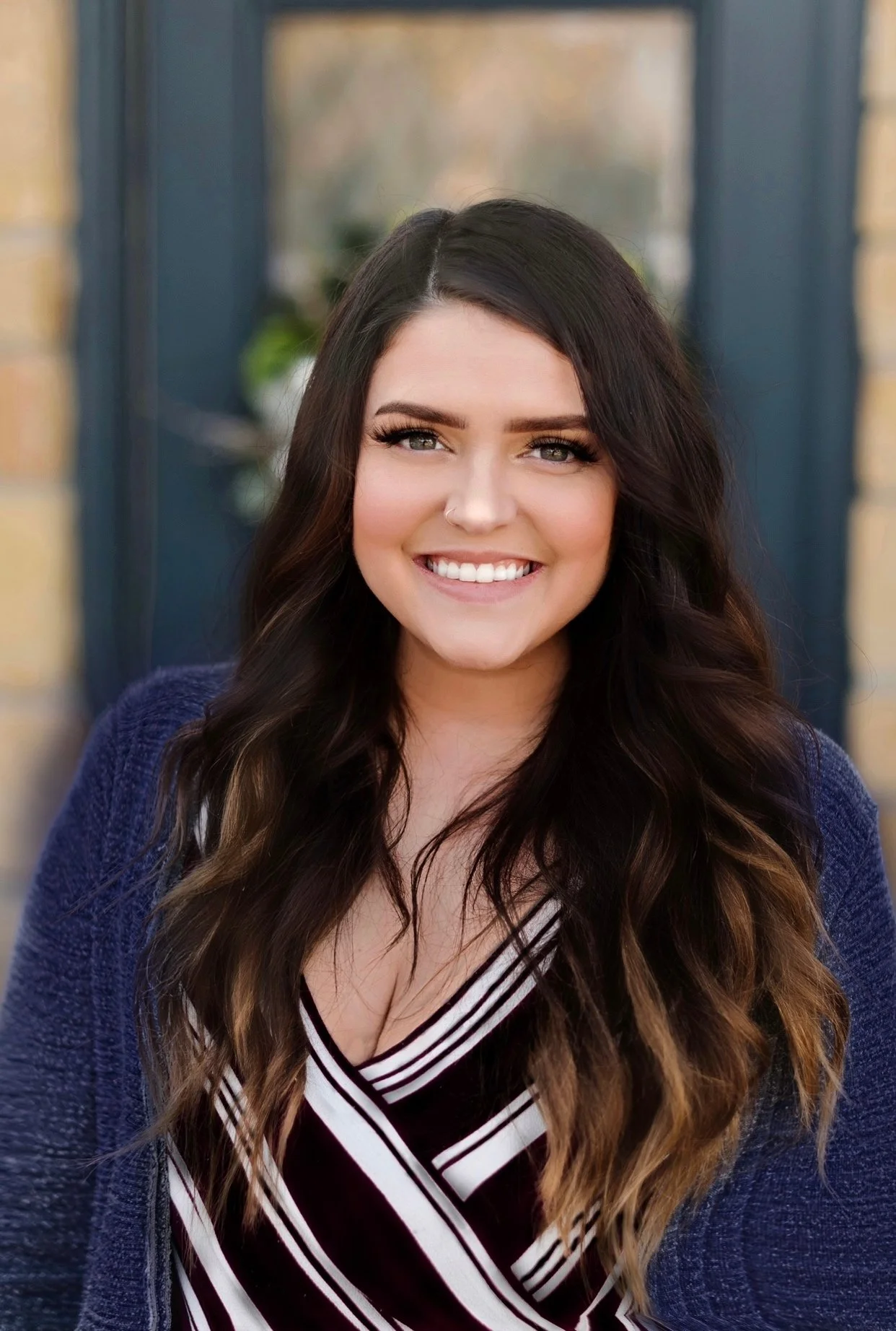 A young woman with long, wavy dark hair, smiling showing her teeth, wearing a navy blue jacket and a black and white striped top.