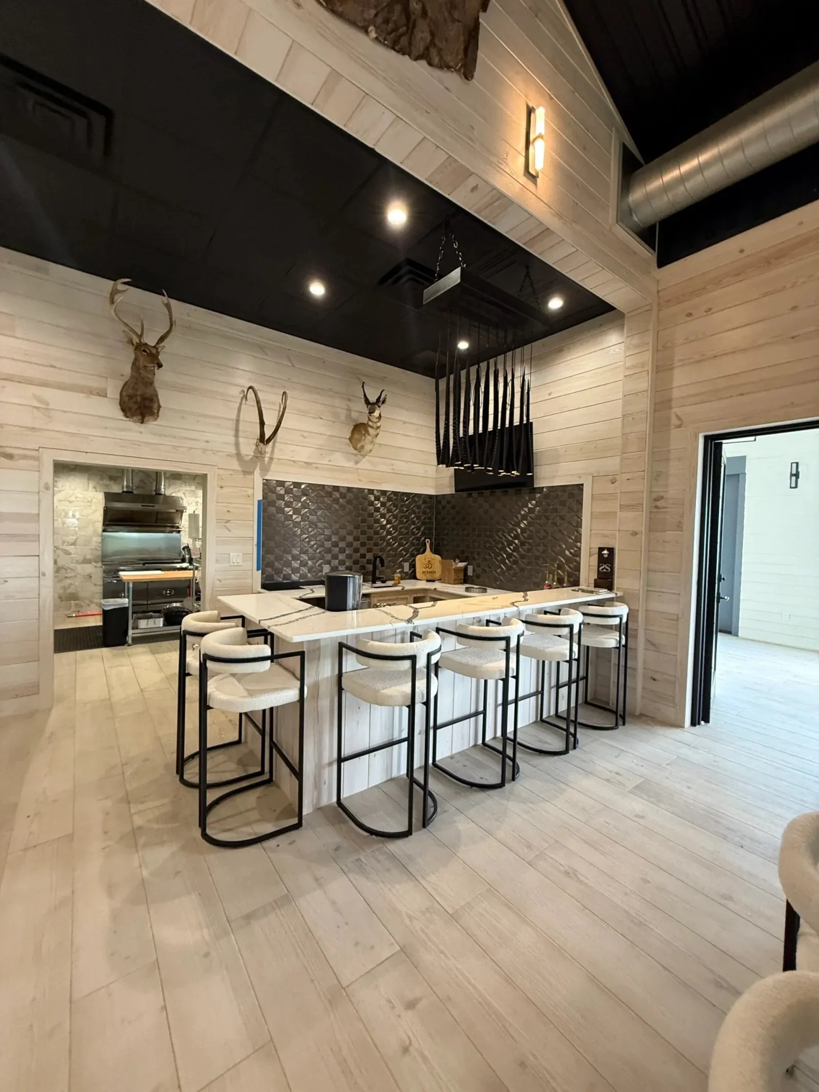 Modern kitchen with a white marble island, six bar stools, wood-paneled walls, mounted deer heads, and a black ceiling with recessed lighting.