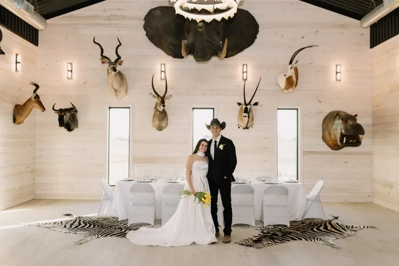 A bride and groom standing together in a decorated room with mounted animal heads on the walls, a zebra rug on the floor, and a zebraskin table runner. The bride has a bouquet of yellow flowers and is in a white wedding dress, while the groom is in a black suit with a black hat.