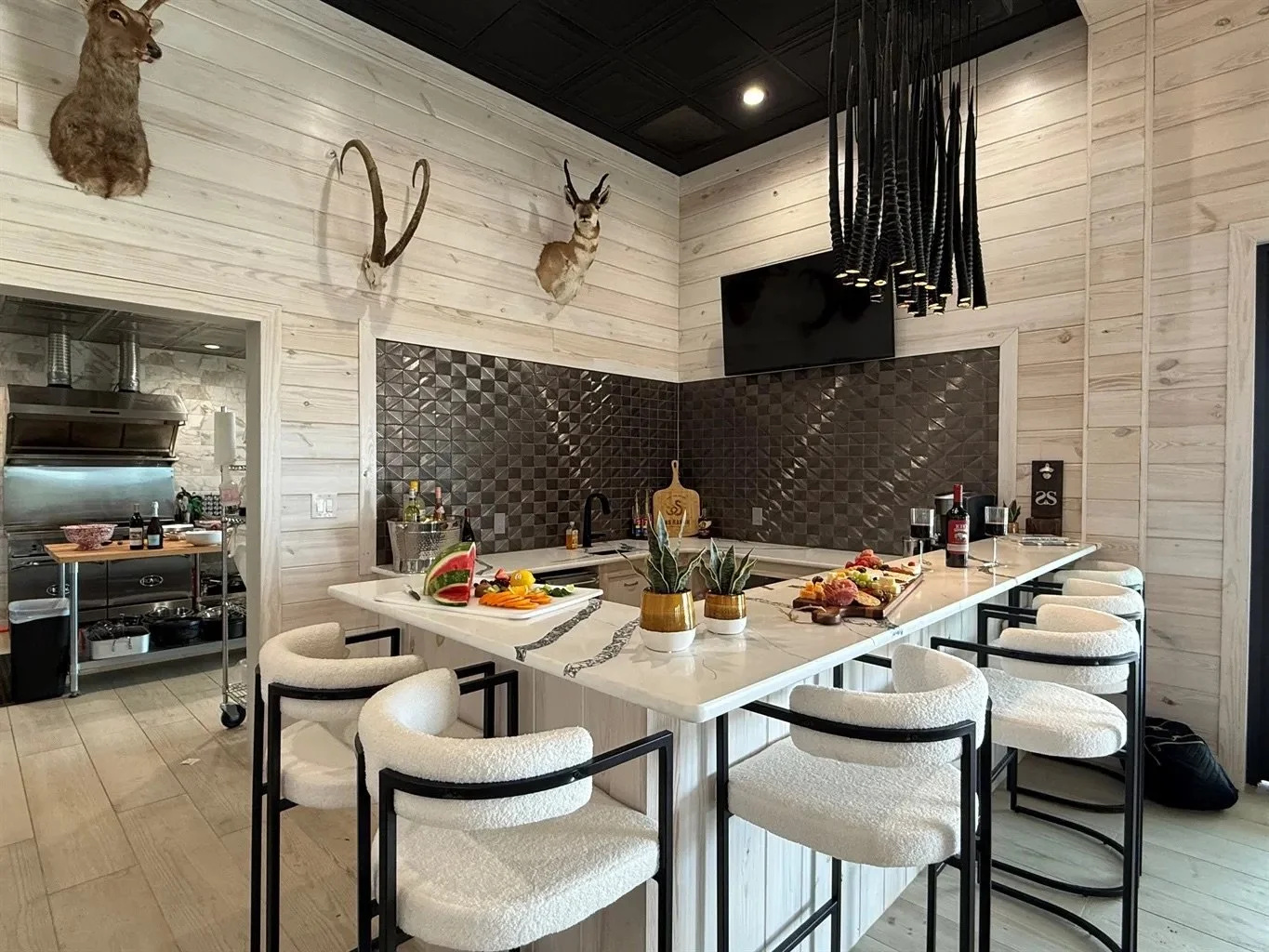 A modern kitchen with white wooden walls, black ceiling, mounted animal skulls and antlers, and a black textured backsplash. An island counter has white chairs, plants, and food items on it.