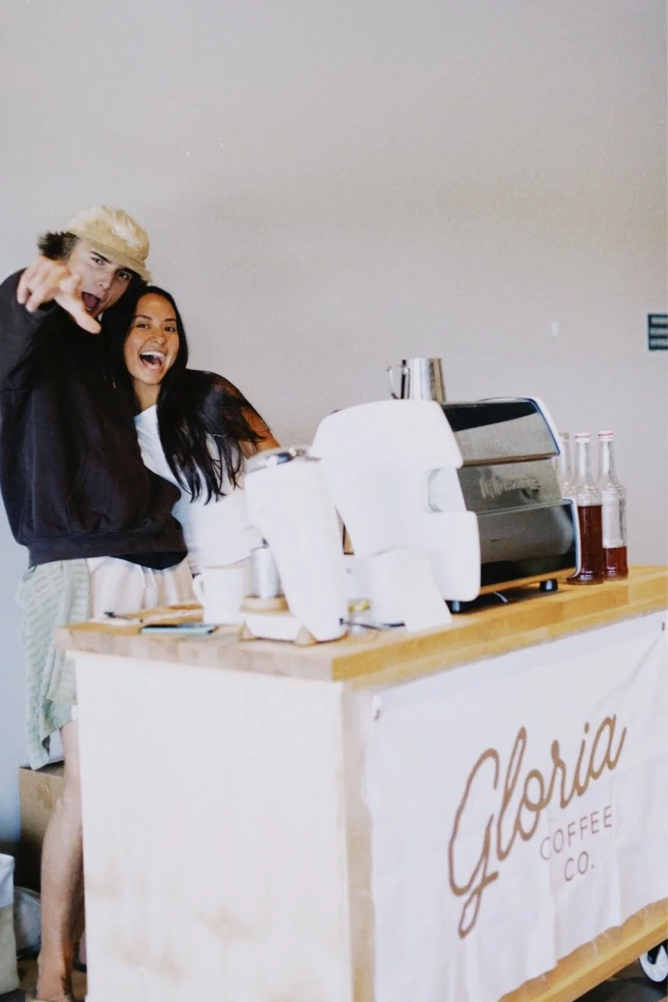 Two people standing behind a coffee cart with a sign that reads "Gloria Coffee Co." They are smiling and appear excited, with one person pointing at the camera. The cart has coffee-making equipment and bottles on it.
