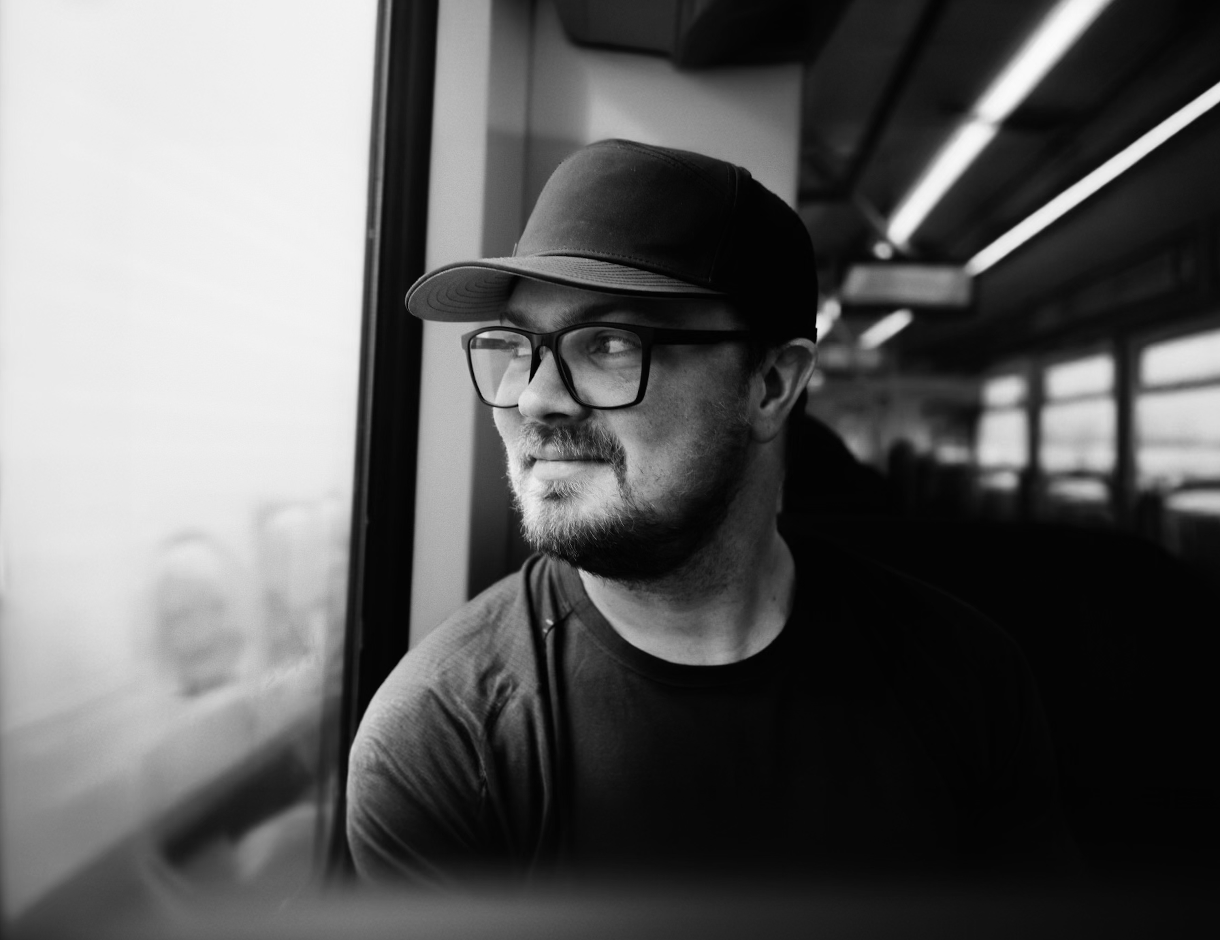 A man with glasses and a beard wearing a baseball cap, looking out of a window on a bus or train.