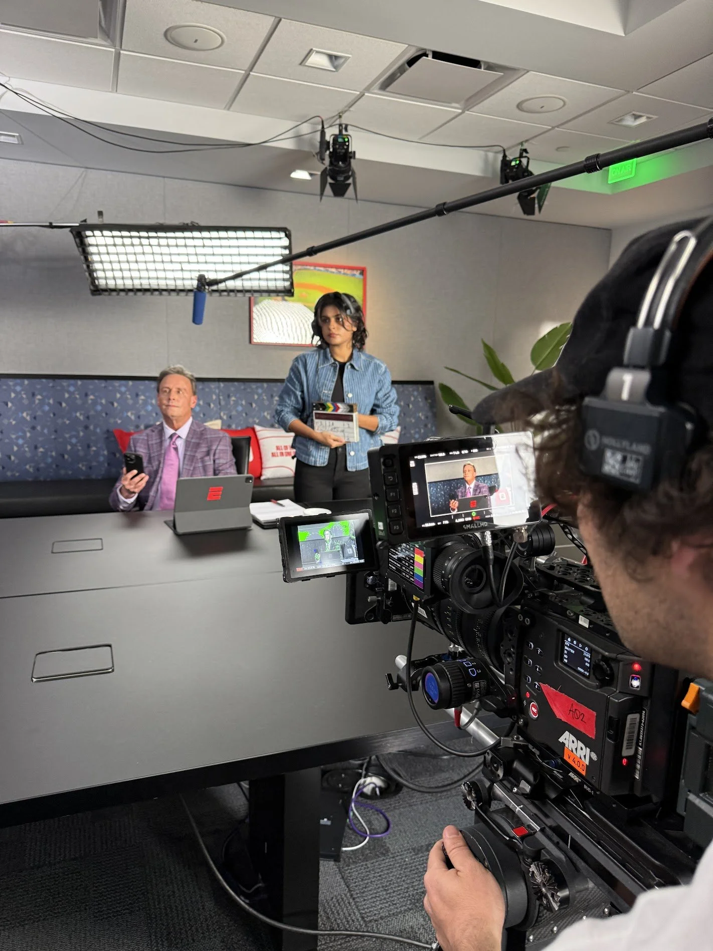 Behind the scenes of a video shoot, a director is filming a woman in a studio. The woman is holding a clapperboard, and a man is sitting at the table looking at his phone. The studio has professional lighting and cameras.