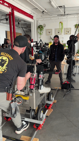 A film crew setting up camera equipment in a studio with white walls and hanging plants.