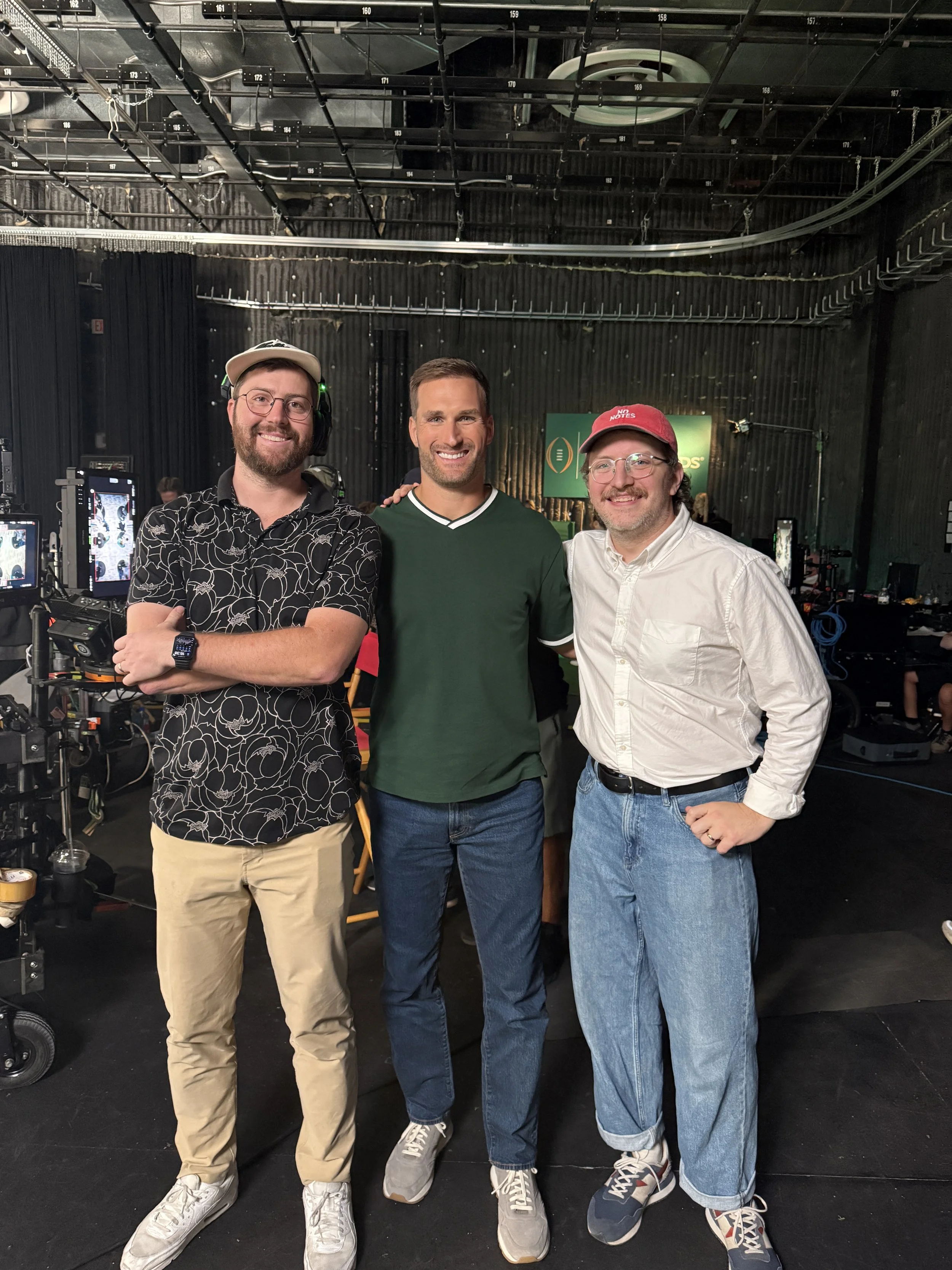 Three men standing together in a dark room with equipment and a green sign in the background. They are smiling and posing for the camera, with the man on the left wearing glasses, a patterned shirt, and beige pants, the man in the middle wearing a green shirt and jeans, and the man on the right wearing glasses, a red cap, and a white shirt with rolled-up sleeves.