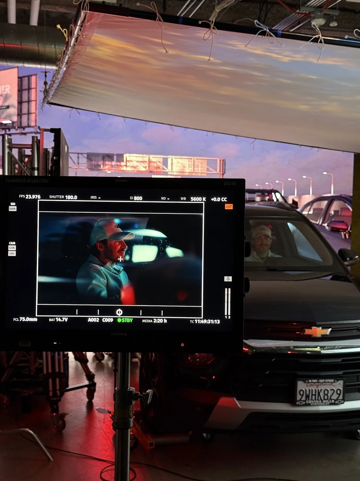 Behind the scenes of a film or photoshoot at a parking lot during sunset. A monitor shows a close-up of a man wearing a baseball cap, sitting in a car, with a reflection of a screen or light in the background. Several parked cars and streetlights are visible outside.