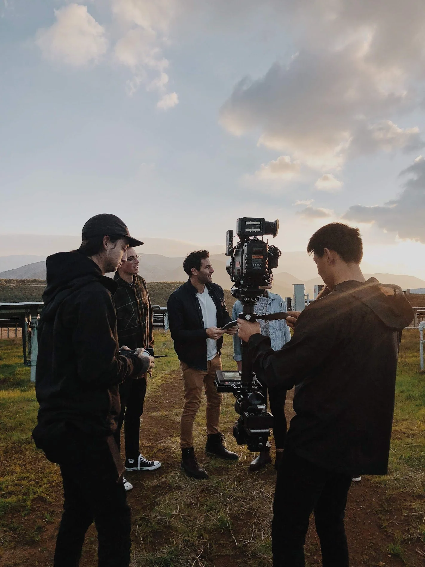 A group of five young men preparing to film outdoors during sunset, with a camera mounted on a tripod and a cloudy sky in the background.