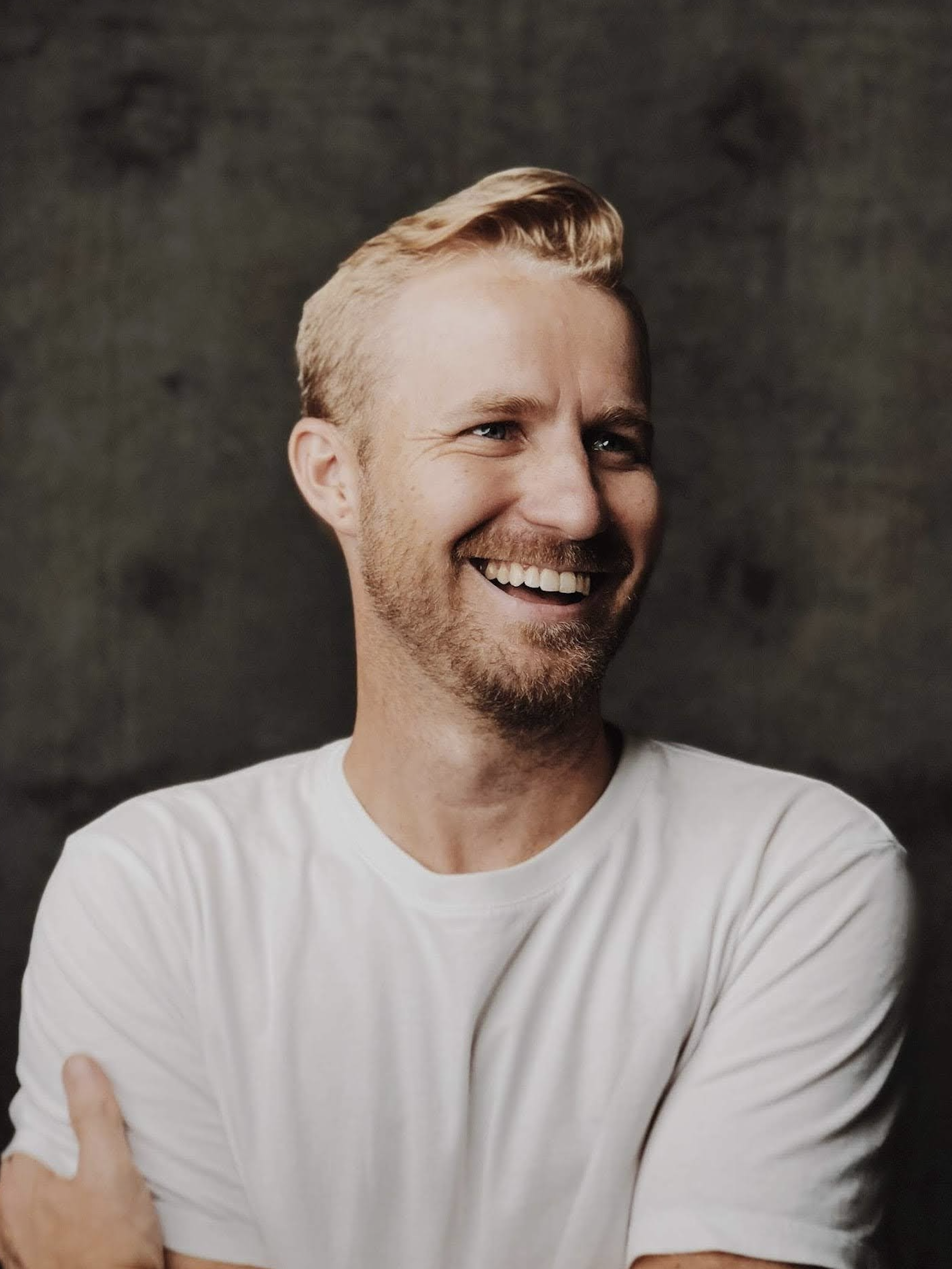 Close-up of a smiling man with blonde hair wearing a white shirt against a dark background.