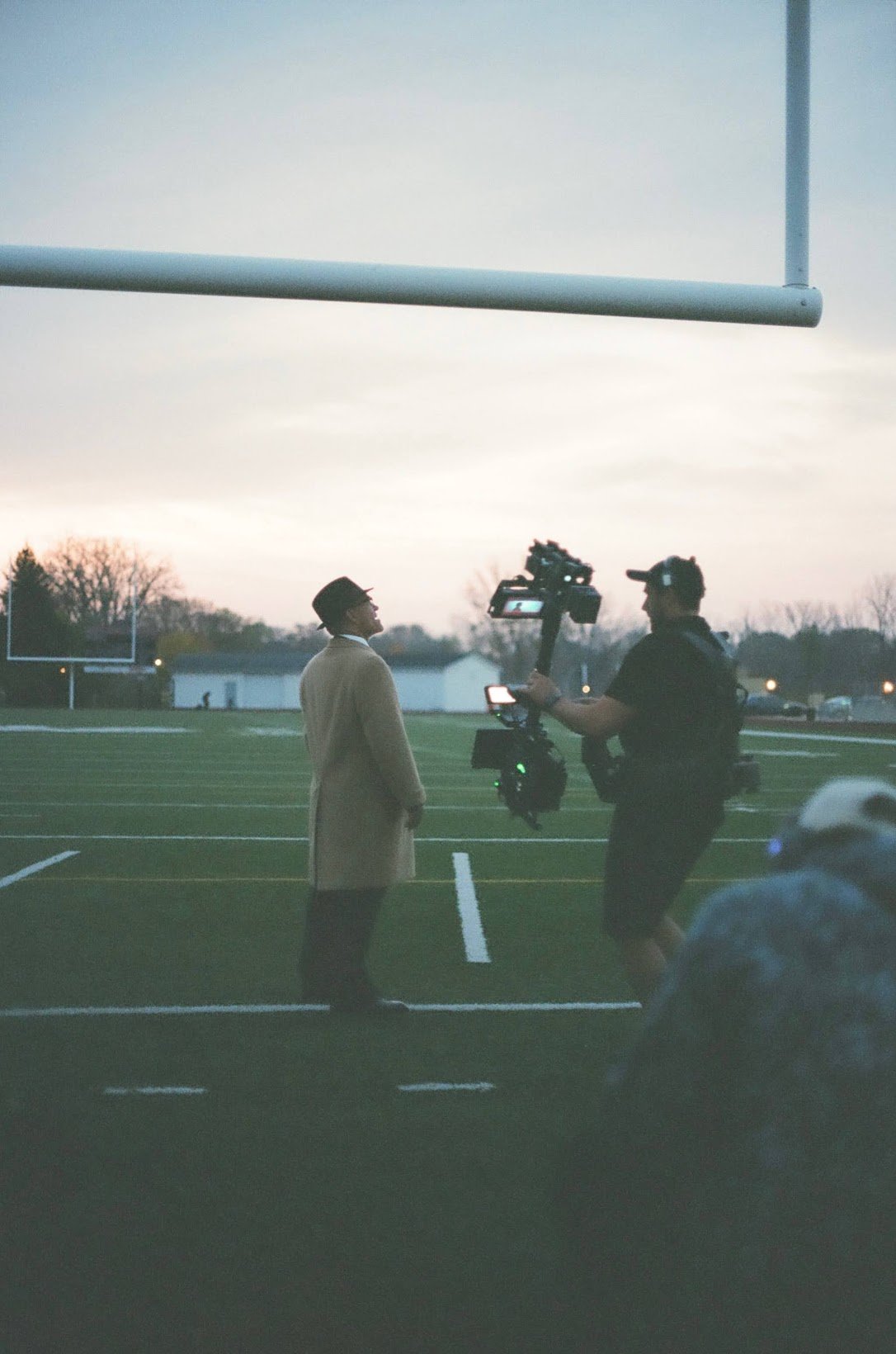 A person in a tan coat and black hat being interviewed by a man with a camera on a football field at sunset.