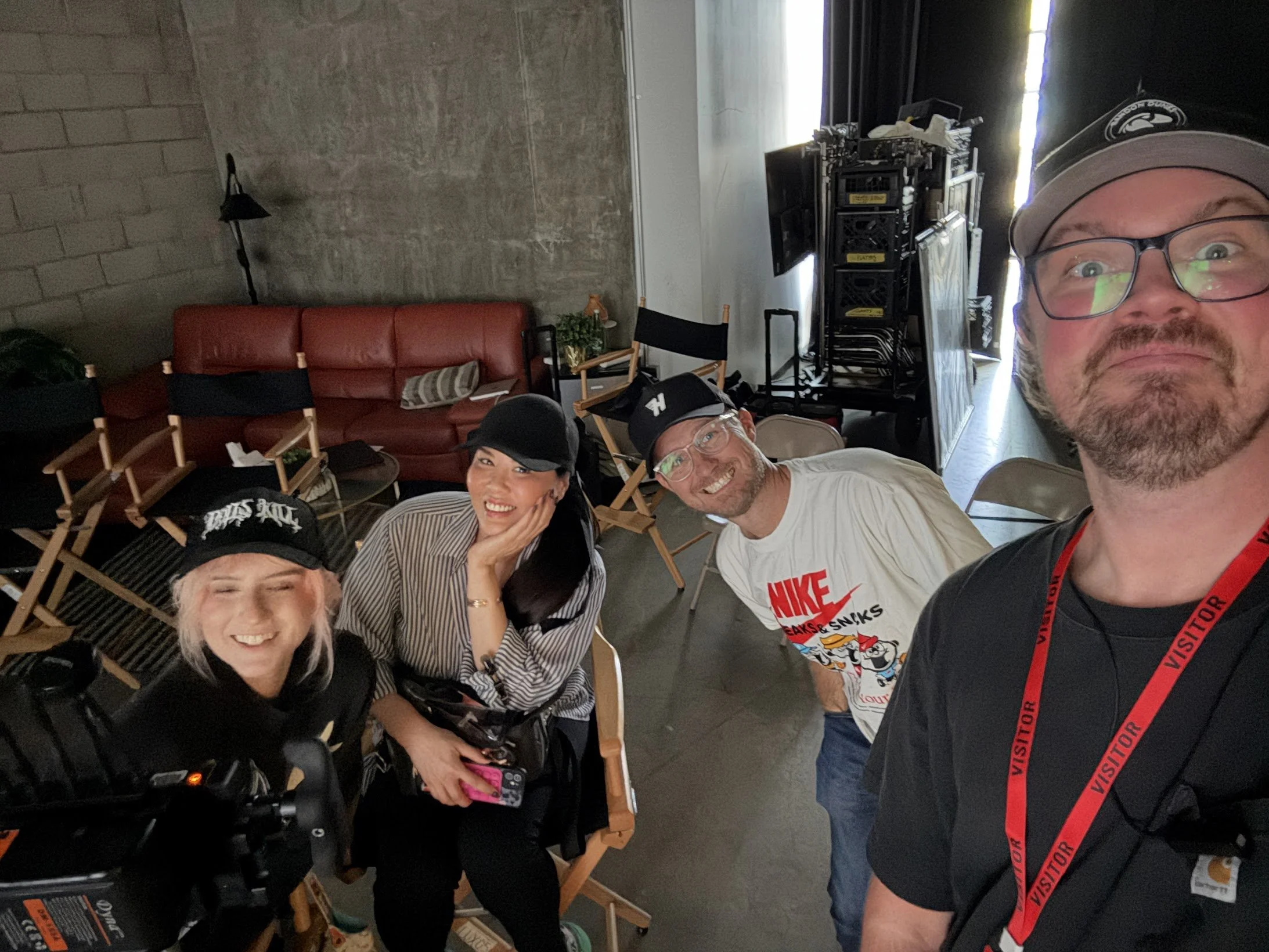 Group of people smiling and taking a selfie in an indoor space with chairs, a red couch, and some equipment or storage boxes in the background.