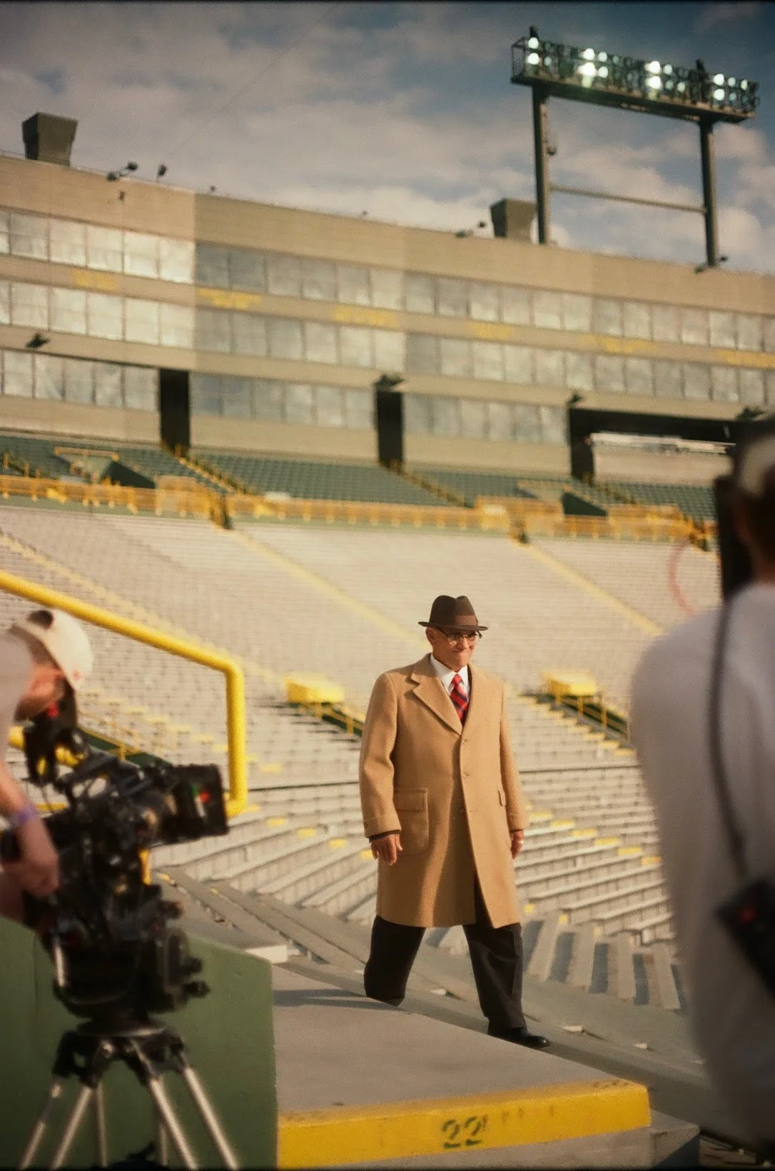 A man wearing a tan trench coat, dark tie, white shirt, glasses, and a brown fedora walking in a baseball stadium with empty stands and a cloudy sky in the background. There are camera crew members nearby.