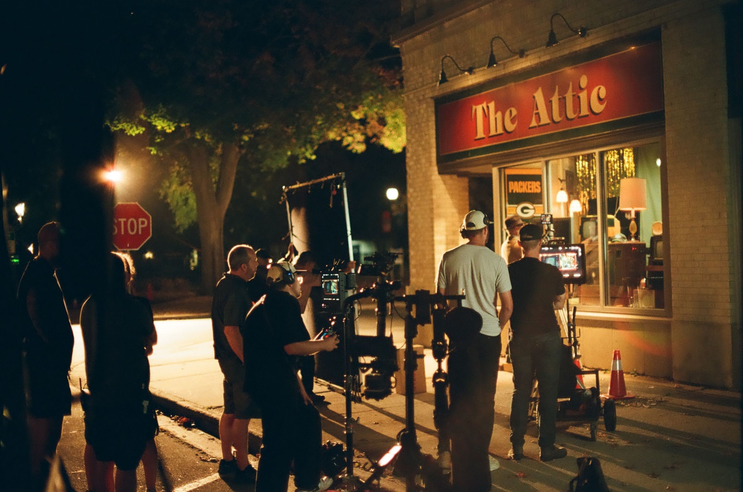 Nighttime scene of a film or video shoot outside a shop called 'The Attic', with crew members and equipment, including cameras and lighting, set up on the sidewalk.